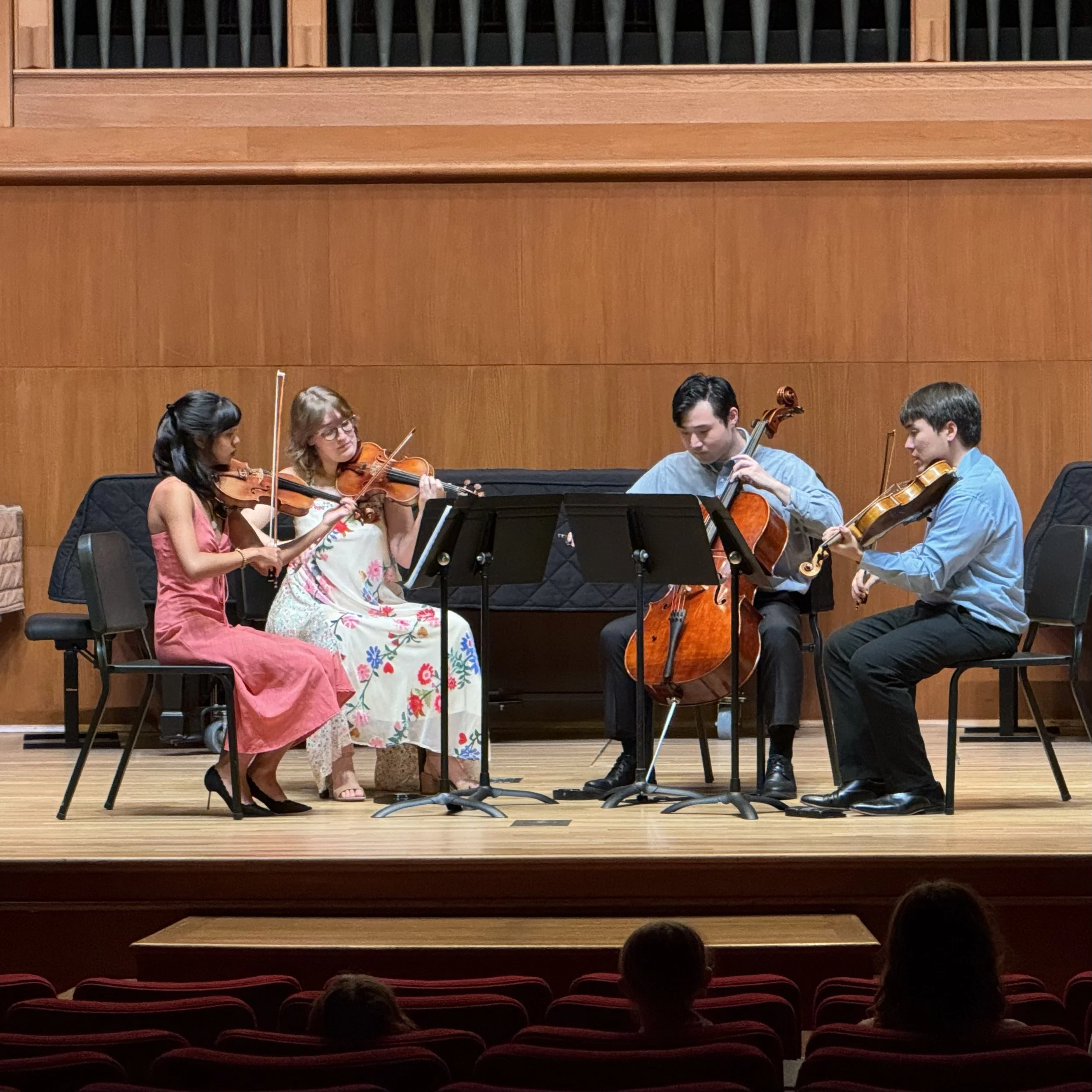 A string quartet performing on stage, including two violinists, a violist, and a cellist, in a concert hall with wooden paneling and an audience in the foreground.