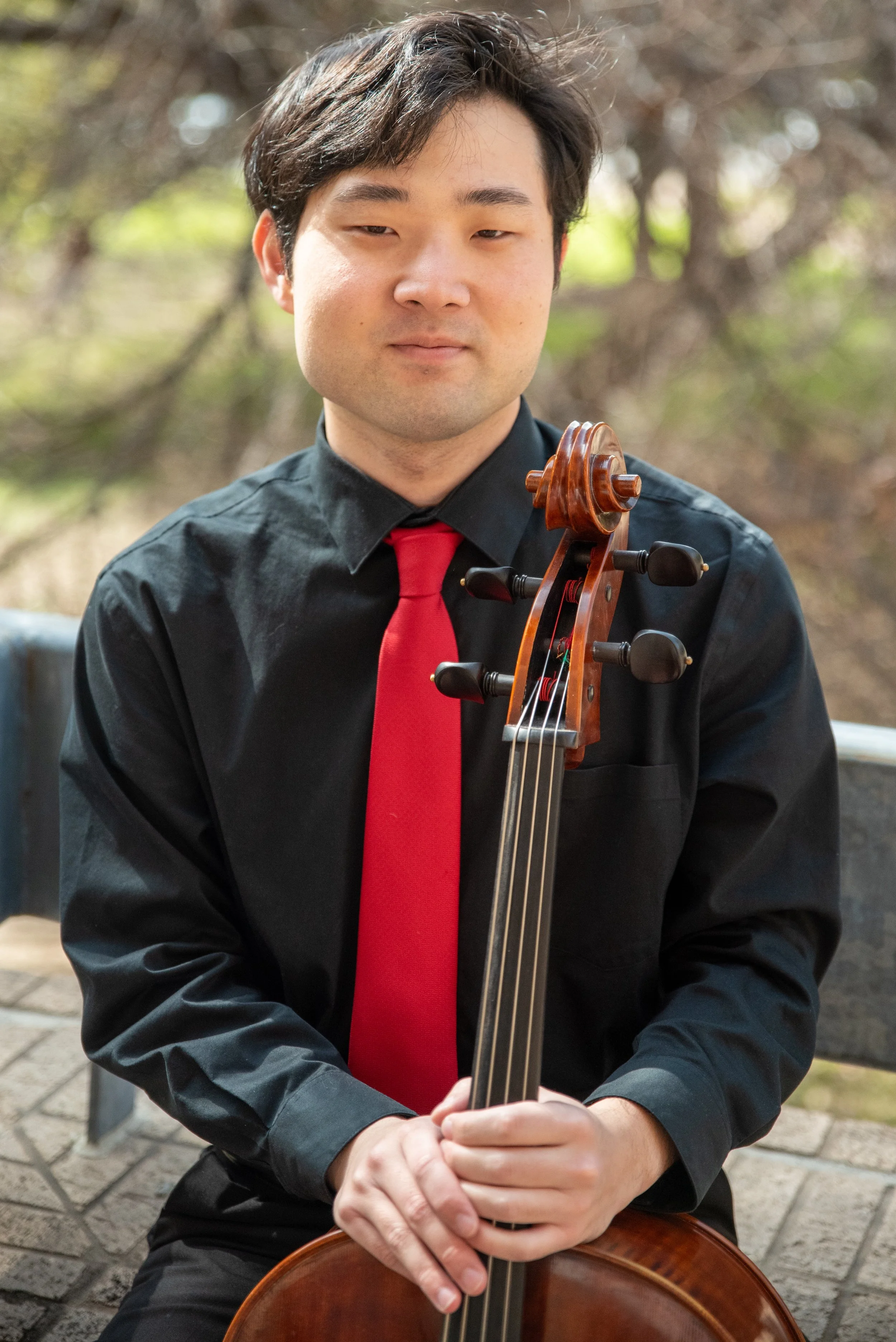 A man dressed in a black shirt and red tie holds a cello, sitting outdoors on a bench with blurred trees in the background.