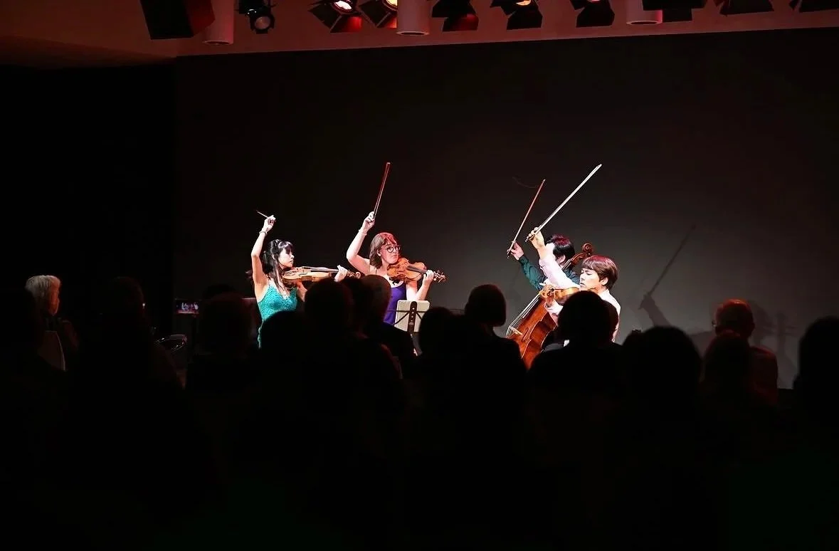 Four musicians during a live performance on stage, with an audience watching in silhouette.