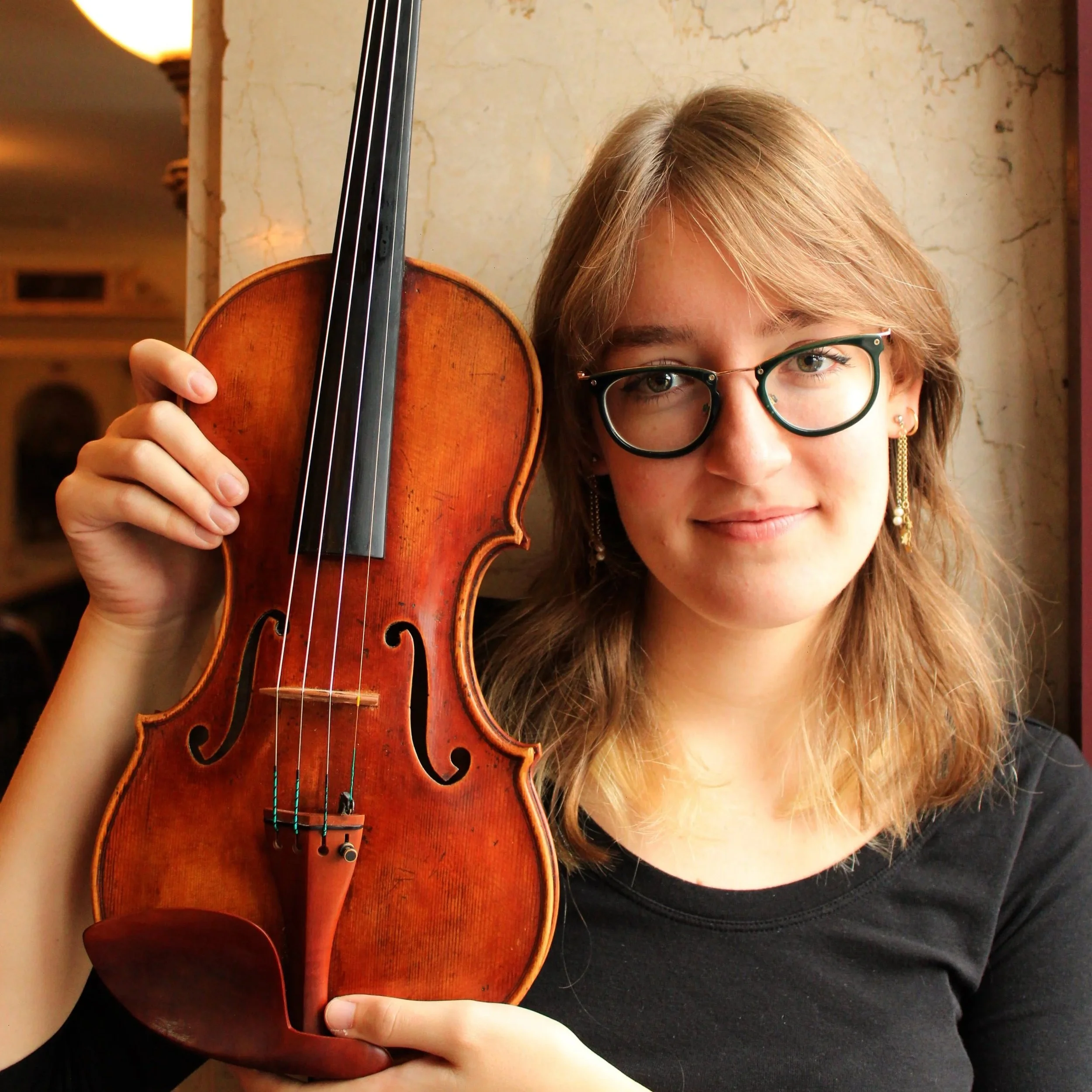 Close-up of a person with light brown hair, wearing glasses, holding a violin near their face, in front of a marble wall with warm lighting.