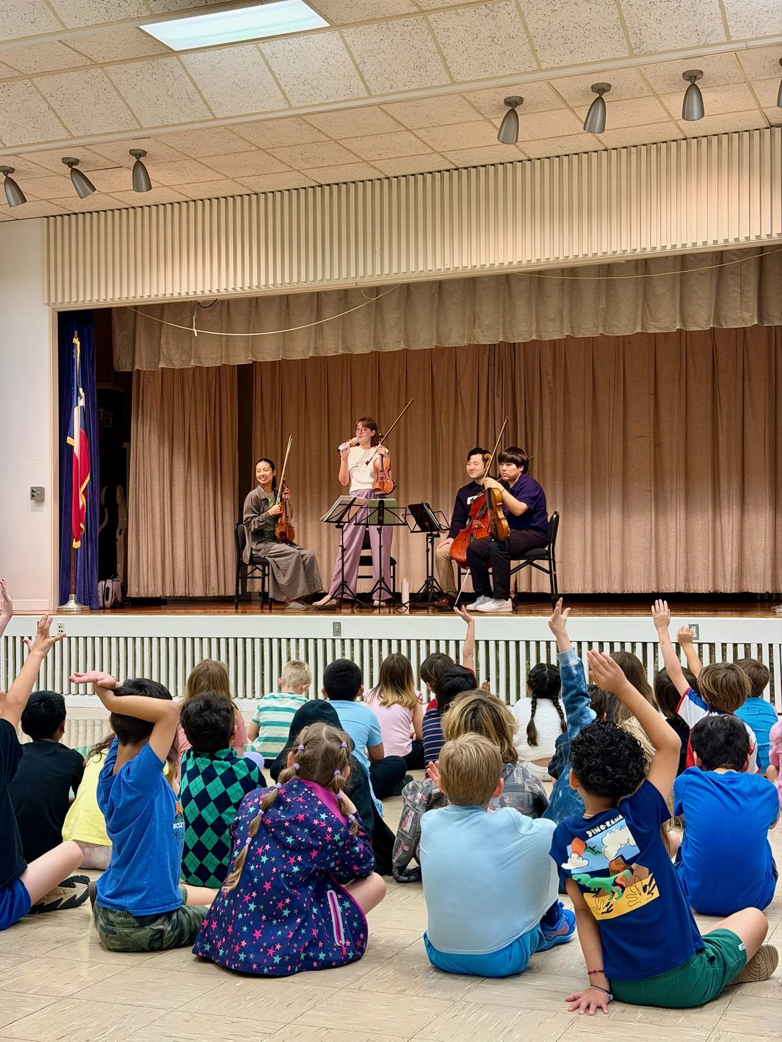 Elementary school children sitting on the floor facing a stage where musicians are performing, with one woman holding a violin and talking into the microphone, and three other musicians holding string instruments.