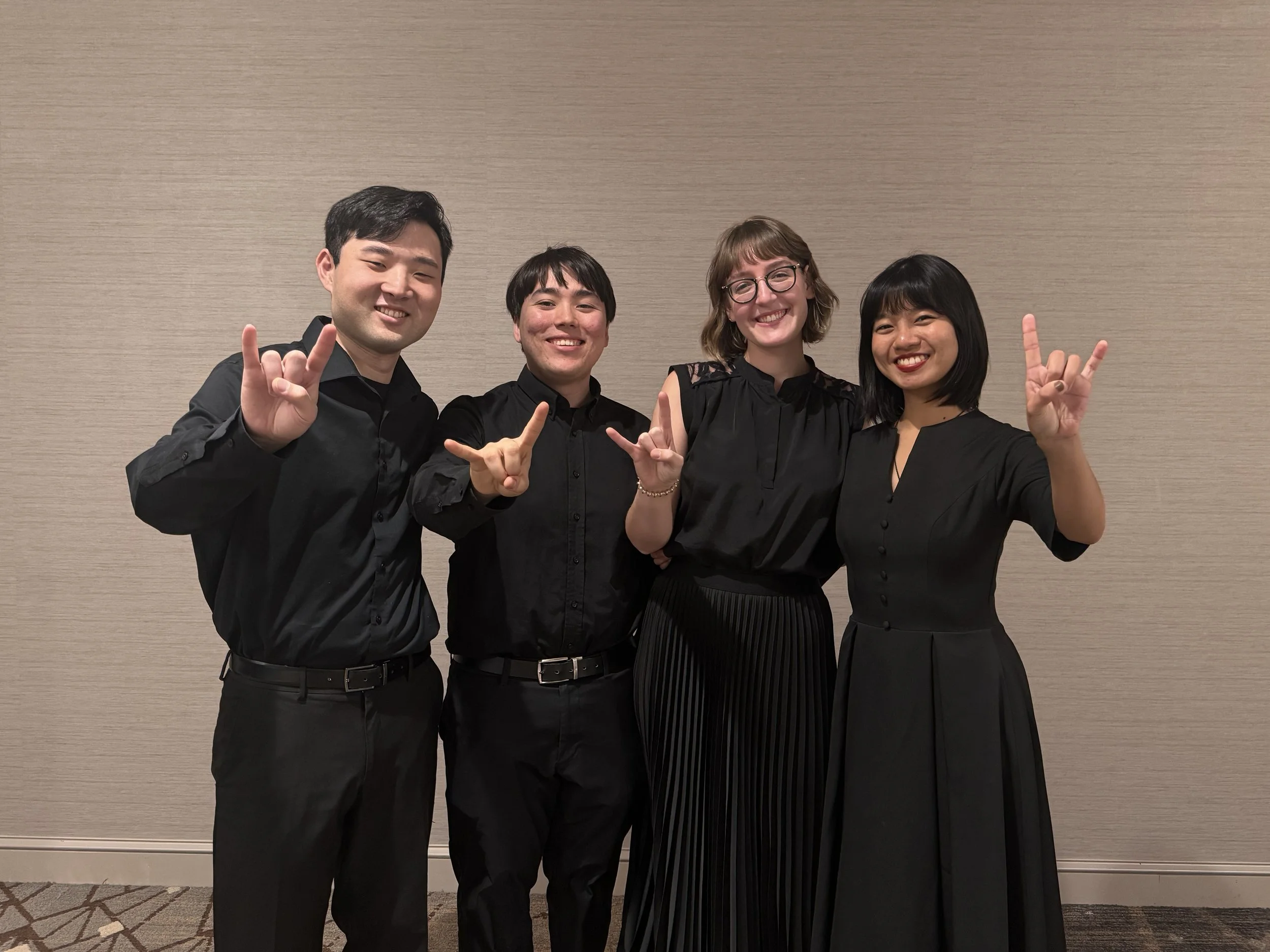 Four musicians smiling and making the University of Texas "Hook 'em" hand sign, dressed in black, standing against a beige wall.
