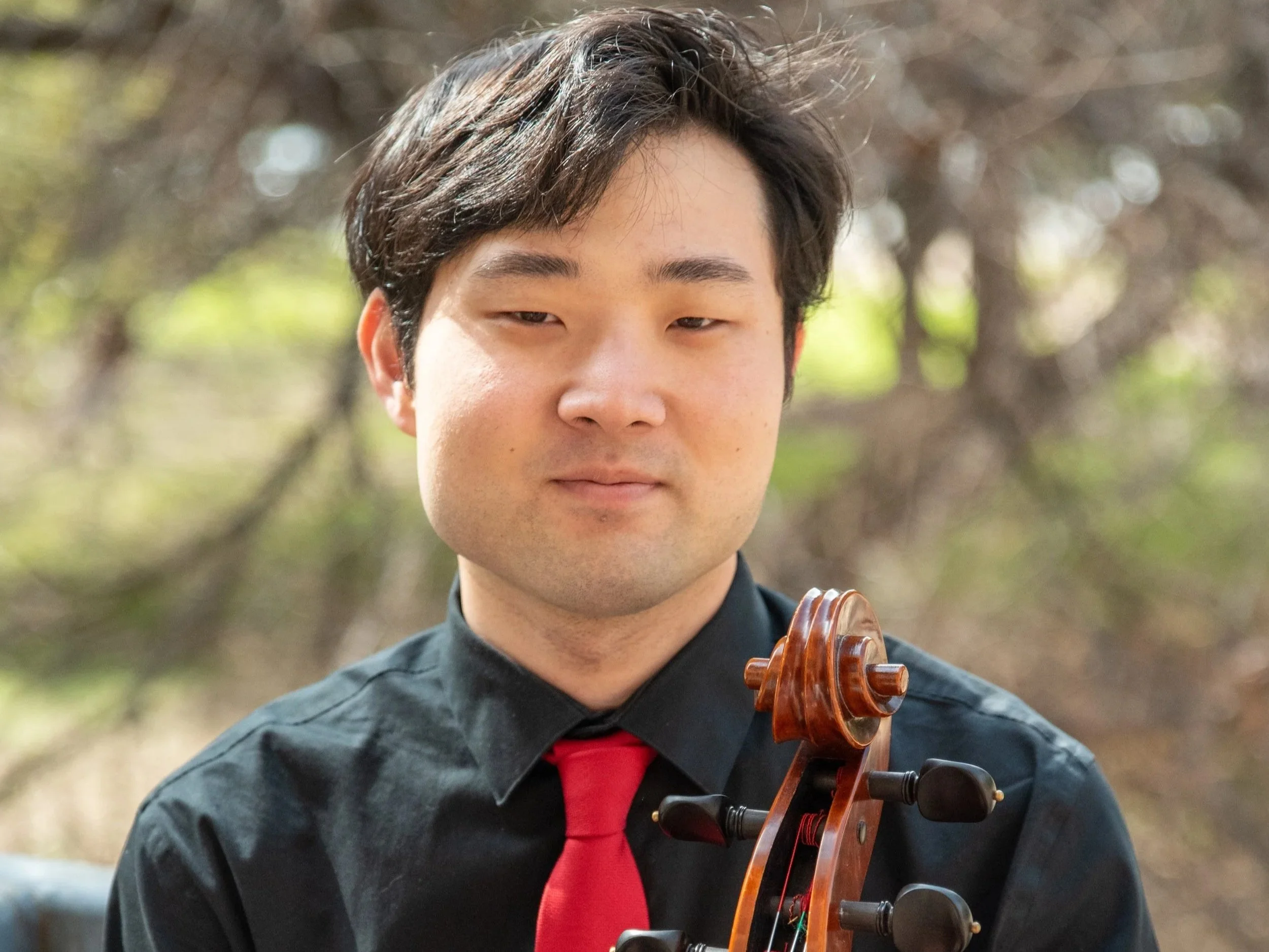 Young man outdoors holding a cello, dressed in a black shirt with a red tie, with trees and blurred foliage in the background.