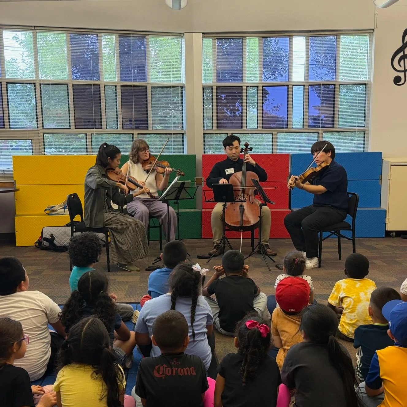 A group of four musicians playing string instruments—two violins, a cello, and a viola—performing in front of an audience of children seated on the floor inside a room with large windows and colorful seating panels behind them.