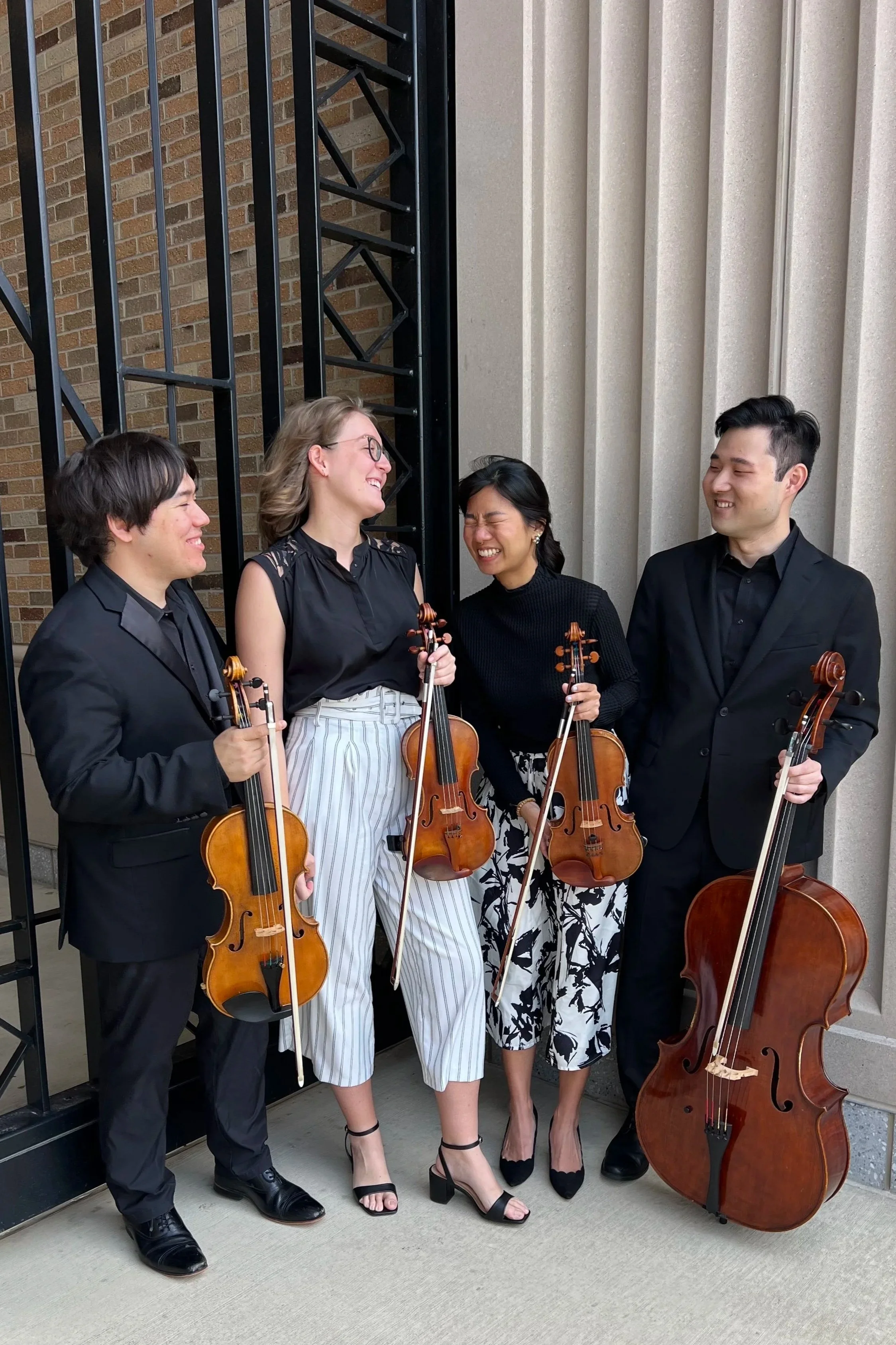 Four musicians stand outside near a brick wall and beige columns. They hold string instruments and are dressed in formal black and white attire, smiling and laughing together.