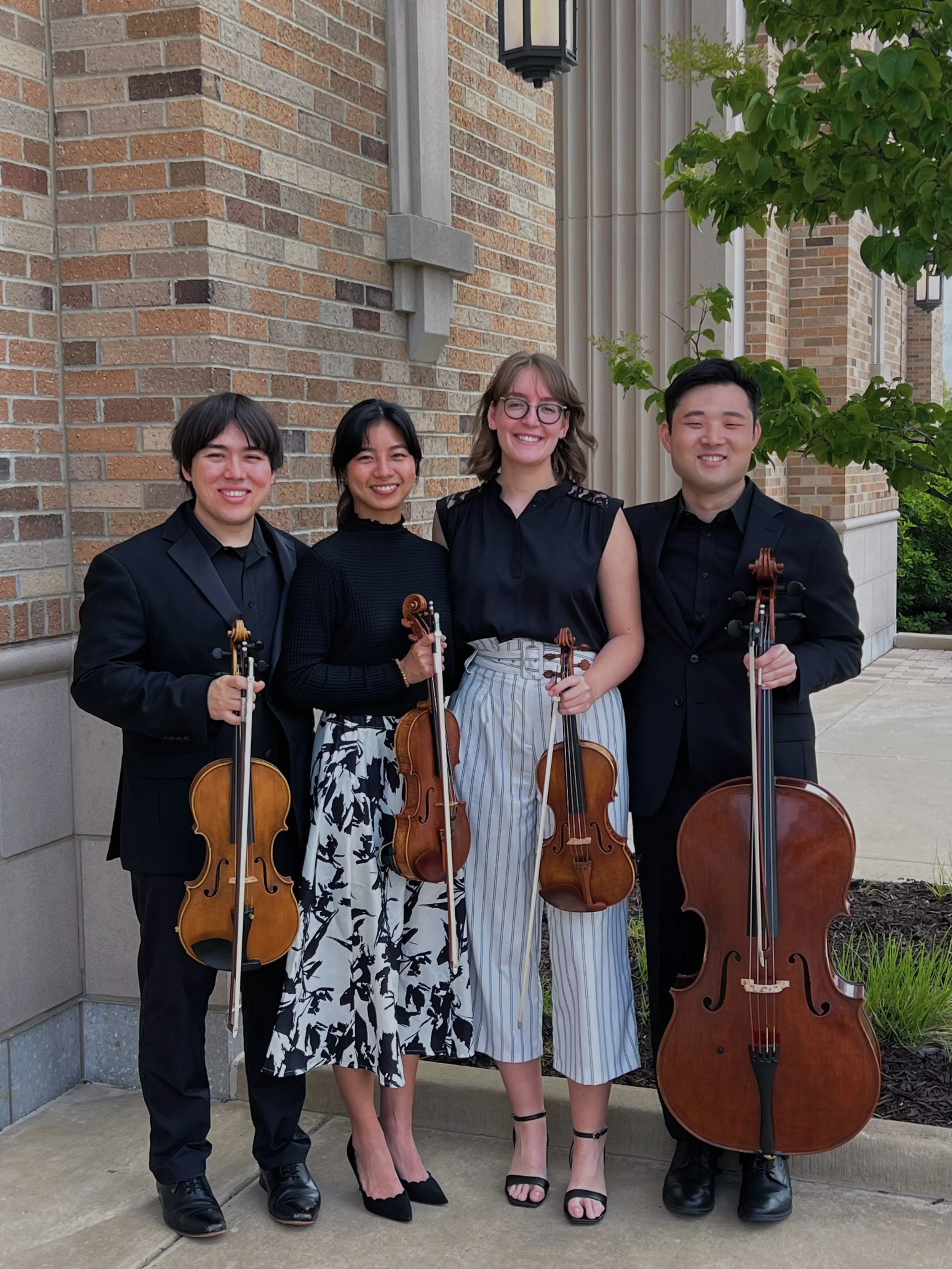 Four young musicians standing outdoors in front of a brick building, holding string instruments. The group includes two men and two women, all dressed in formal black attire, with leafy trees visible in the background.