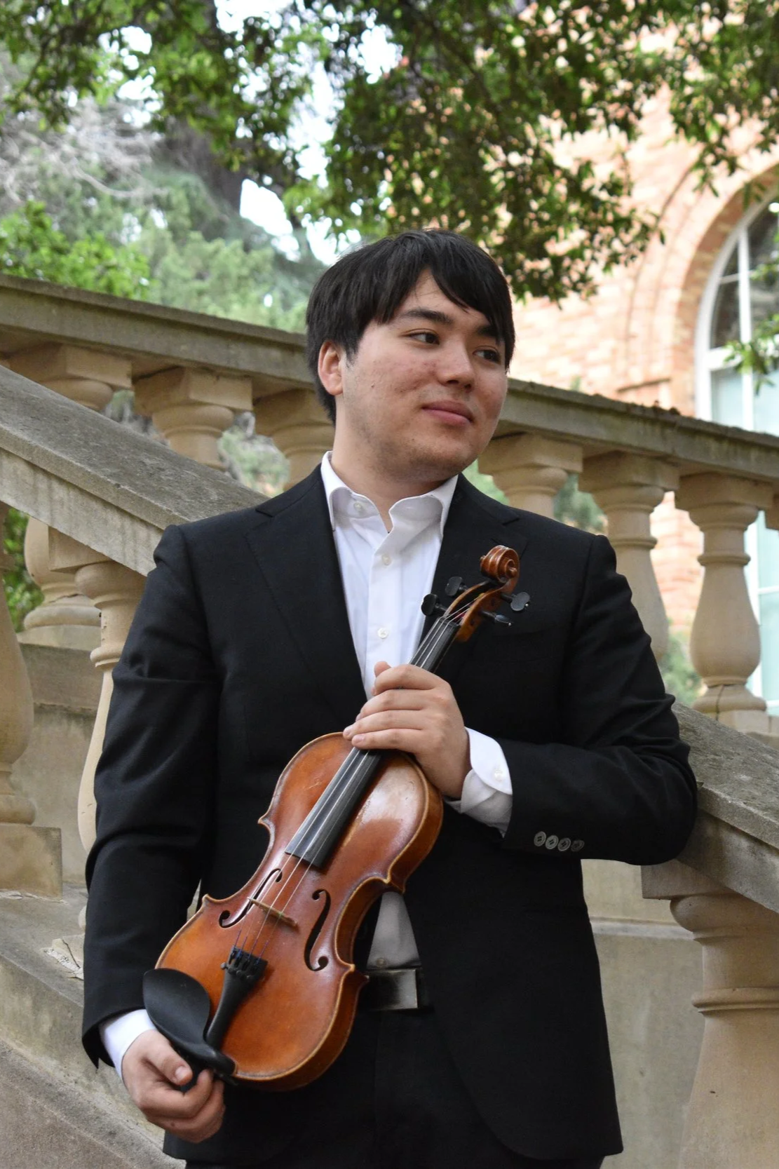 A young man in a black suit holding a violin and bow, standing outdoors near stone stairs and a railing, with trees and a brick building in the background.