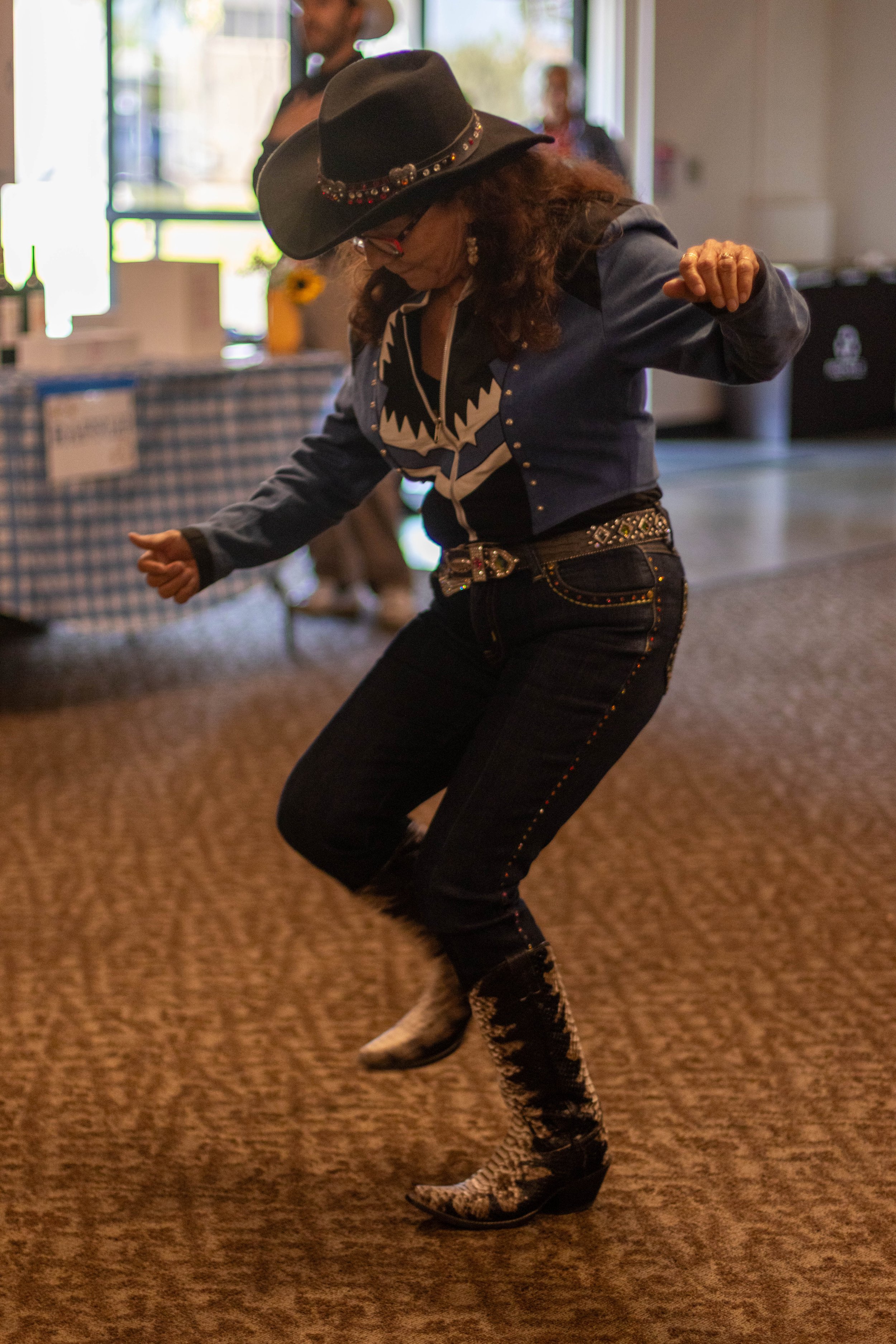 A woman wearing a black cowboy hat with decorative beads, a denim jacket with a graphic design, dark jeans with colorful stitching, and Western-style boots, dancing on a brown carpeted floor indoors with other people and tables in the background.