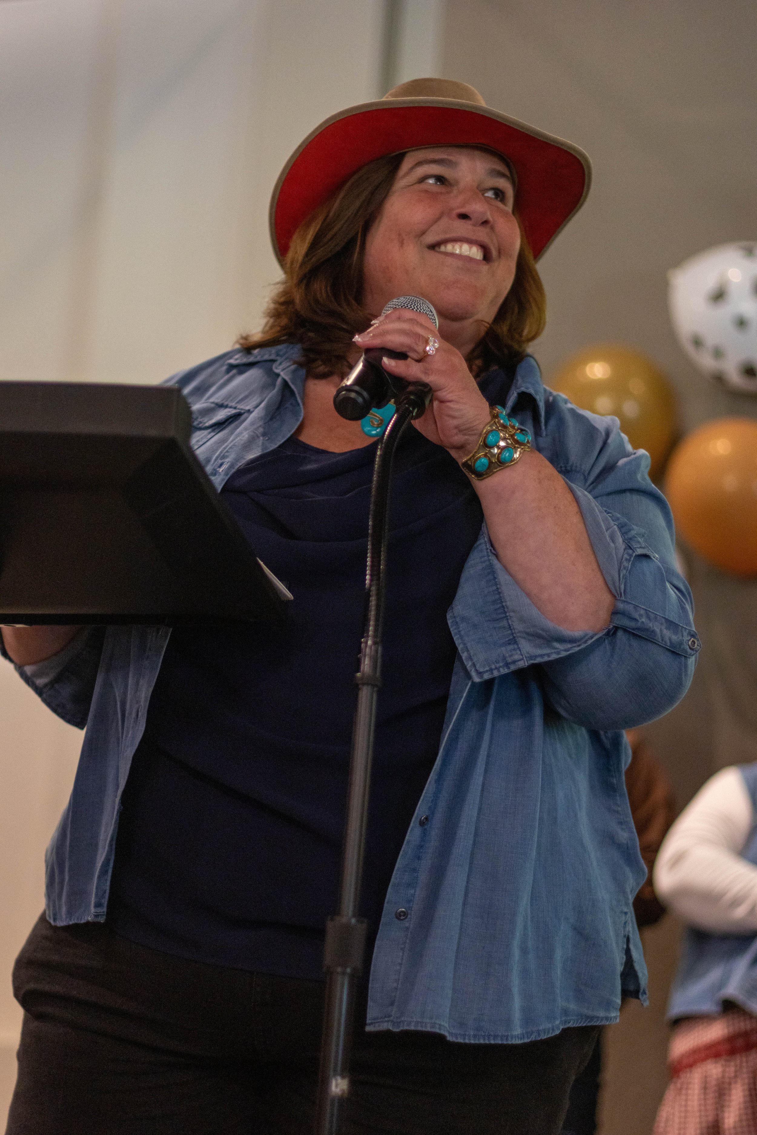 A woman smiling and holding a microphone, wearing a cowboy hat, denim shirt, and various turquoise jewelry, in a room with balloons.