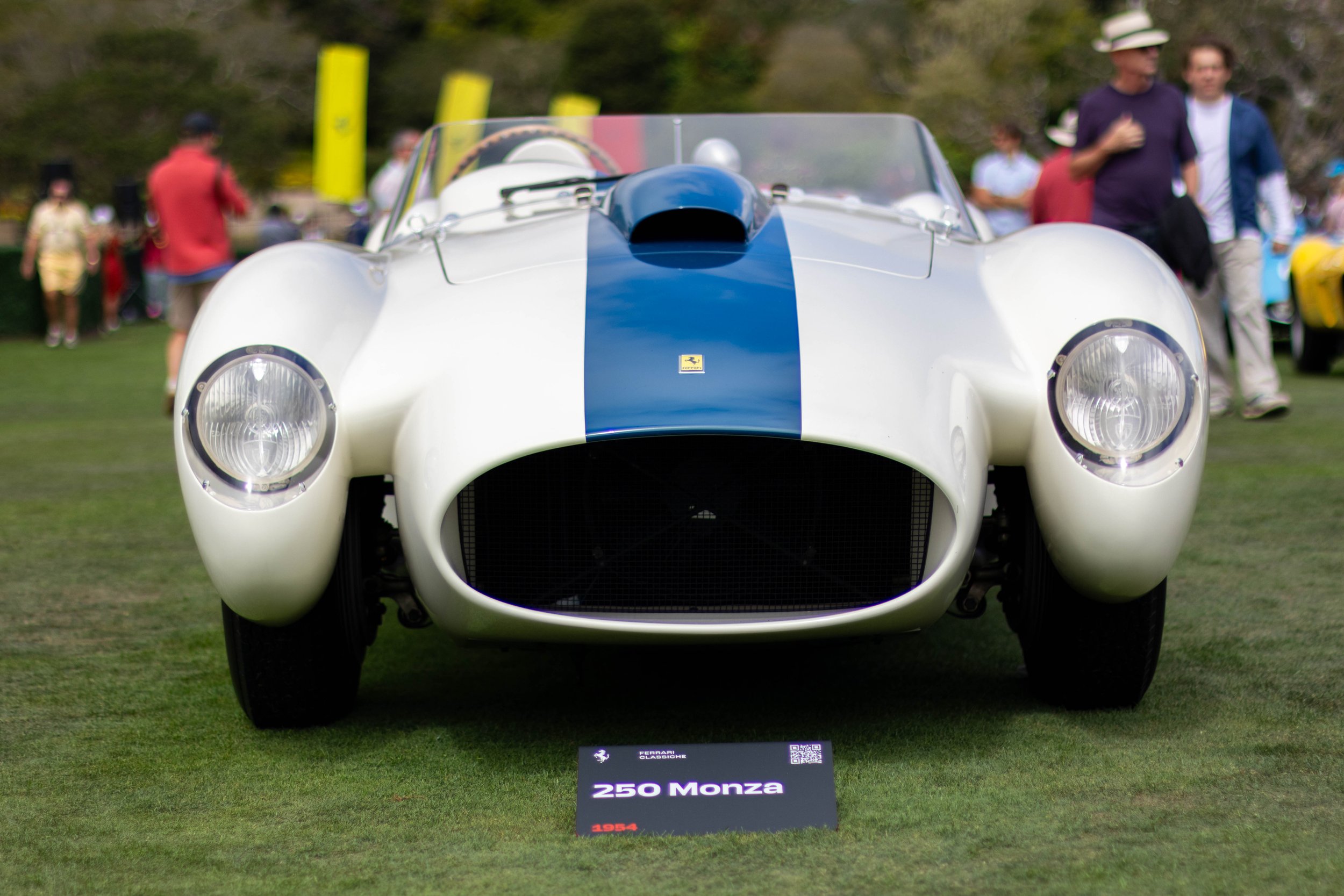 A vintage white race car with a blue stripe on the hood, displayed outdoors at an event with people in the background. The car has round headlights and a low, aerodynamic design. A sign in front indicates it is a 1954 Ferrari 250 Monza.