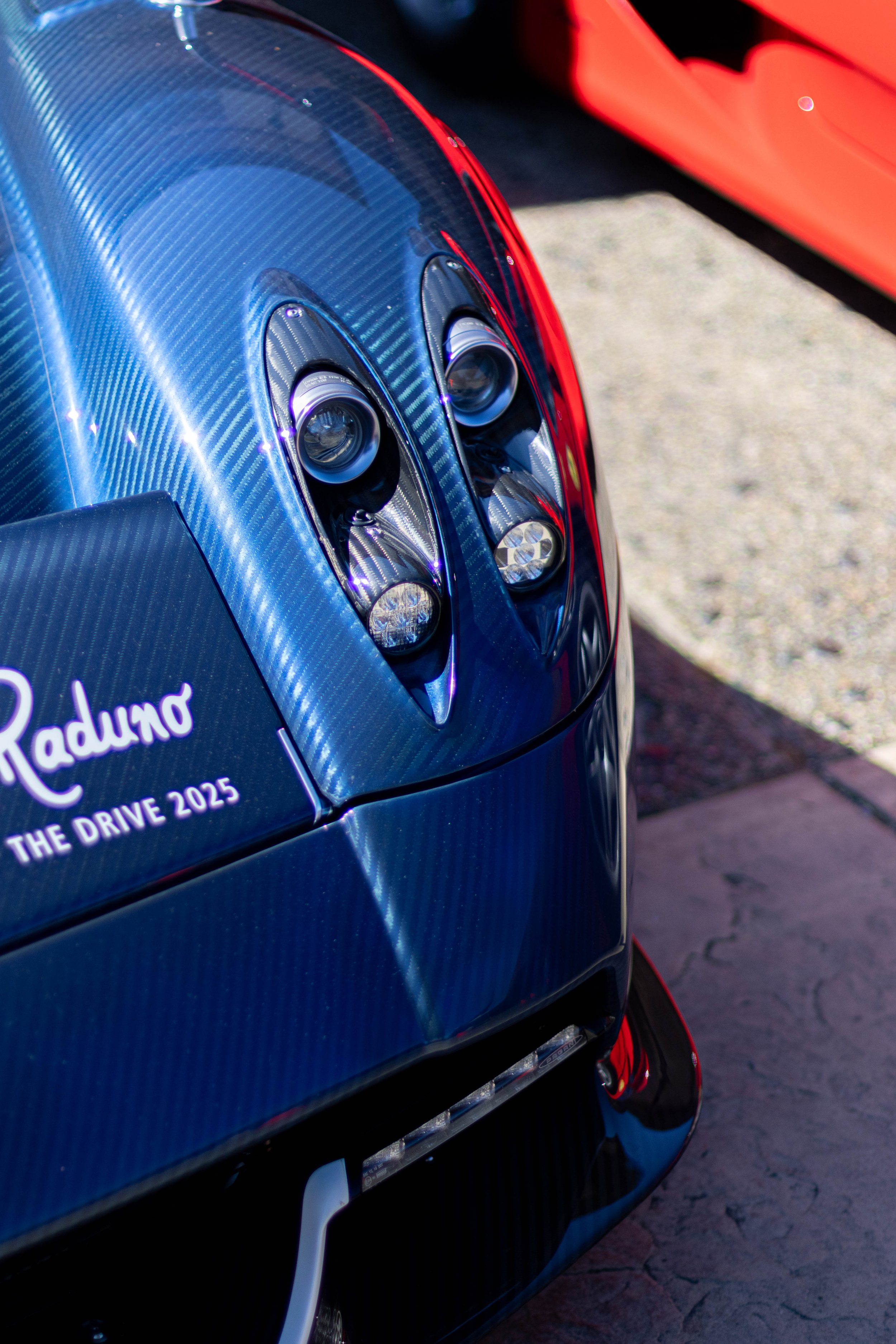 Close-up of a blue sports car with a textured carbon fiber body and sleek headlight design, parked on a paved surface with part of a red car visible in the background.
