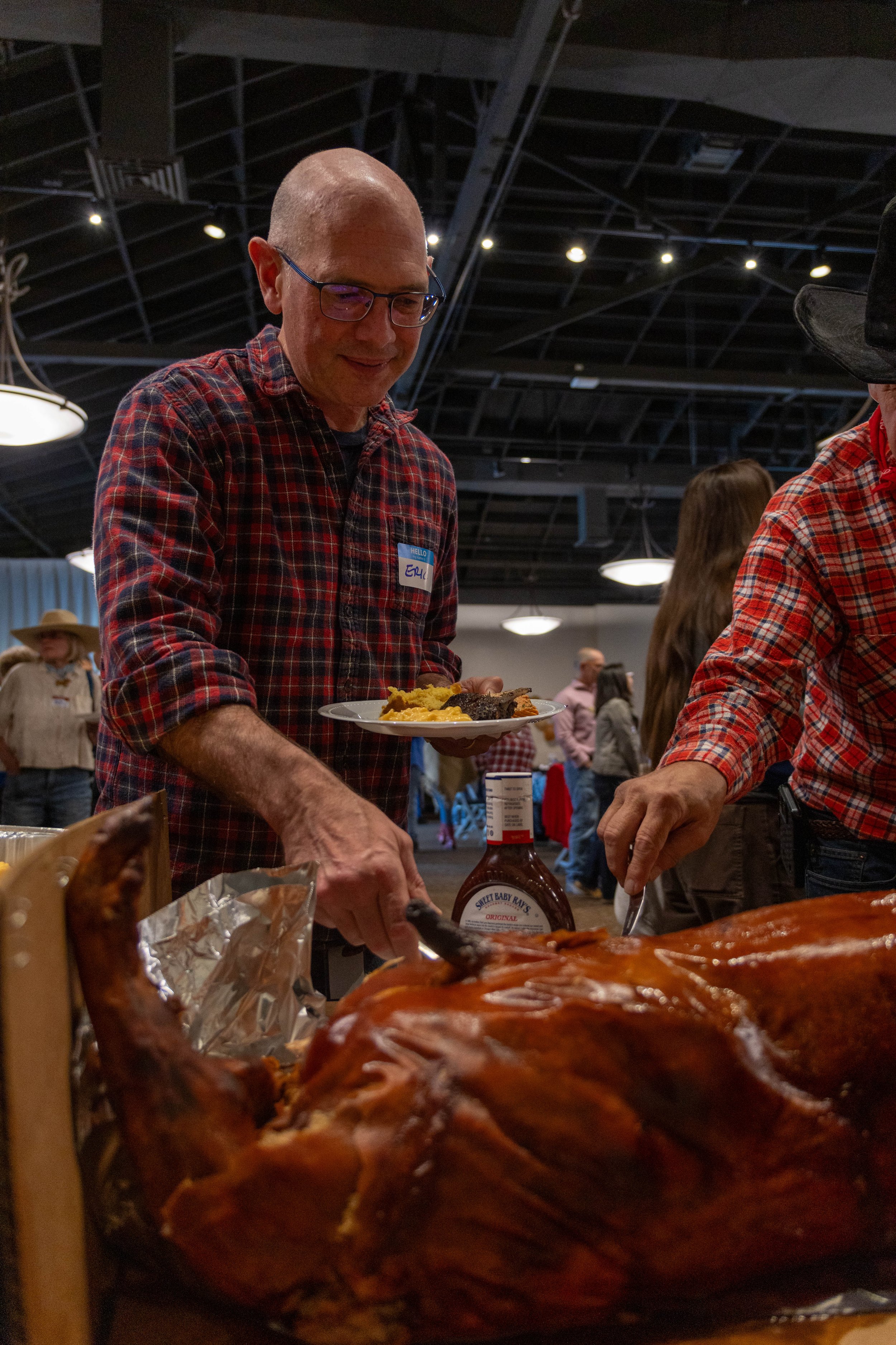 A man with glasses in a plaid shirt holding a plate of food at a buffet, with a large roasted turkey in the foreground, in a busy indoor setting.