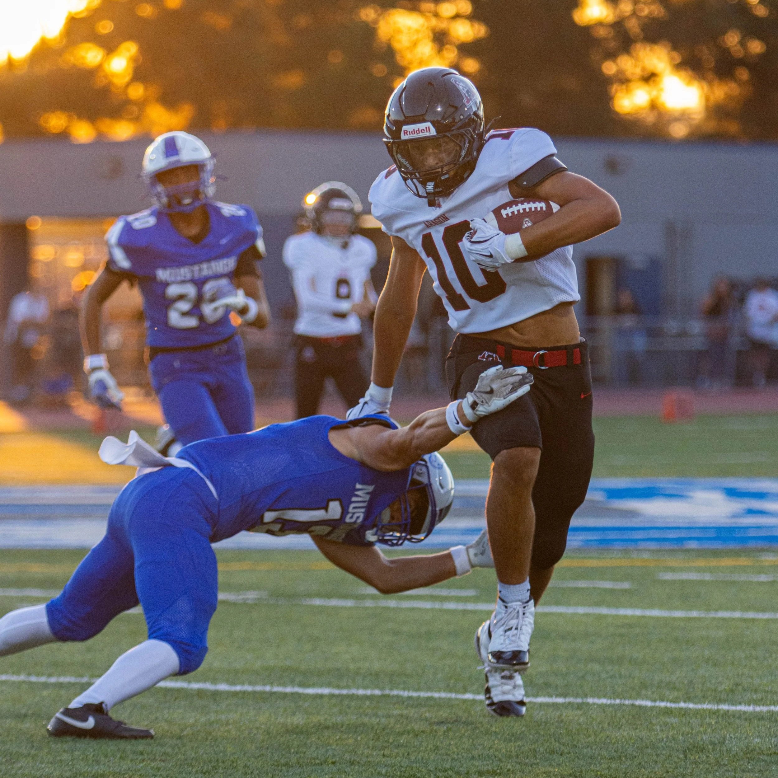 American football game during sunset with players in action, player in white jersey holding football, player in blue jersey on the ground, others observing in the background on a football field.