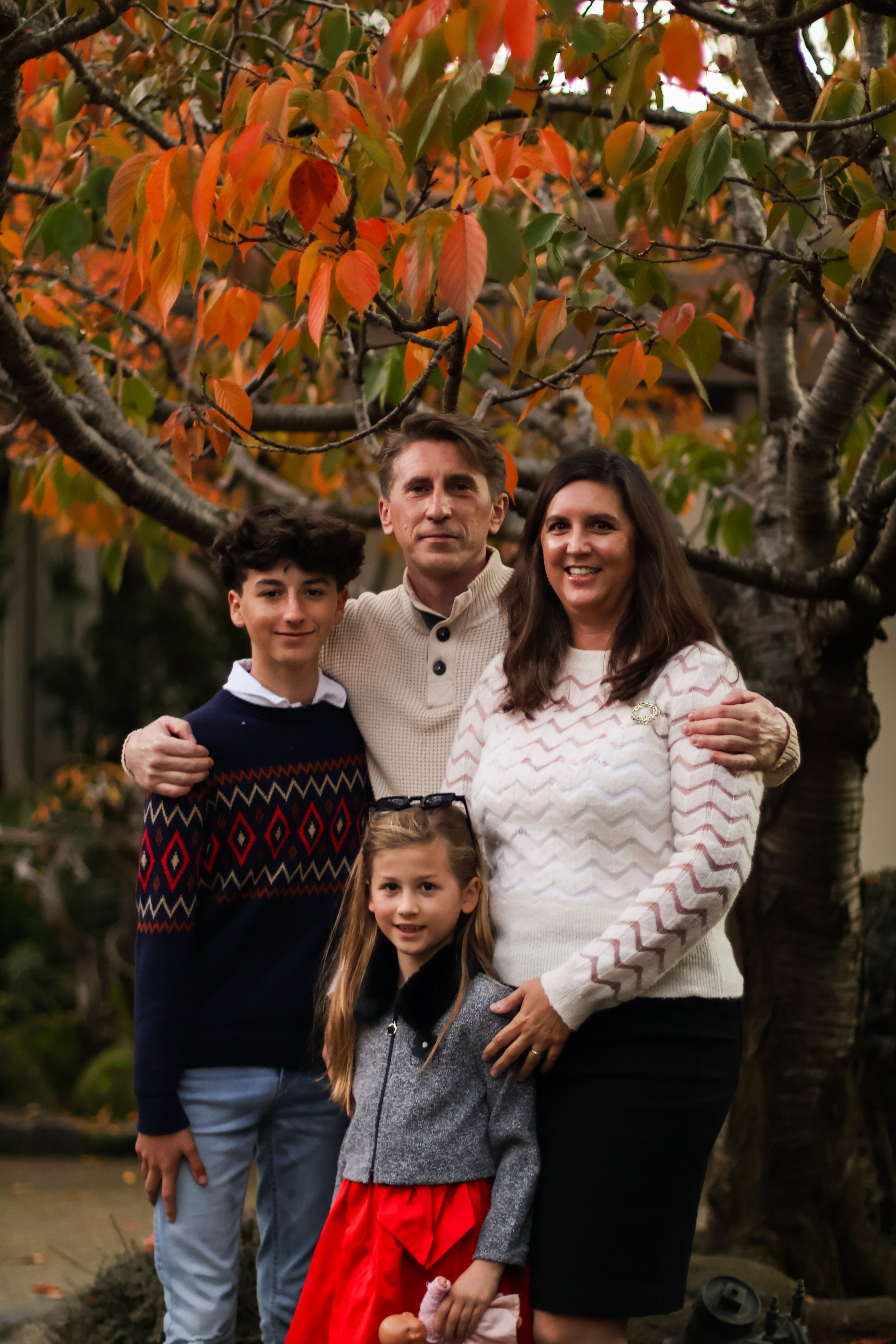 A family of five standing outdoors in front of a tree with orange and green leaves, smiling at the camera.