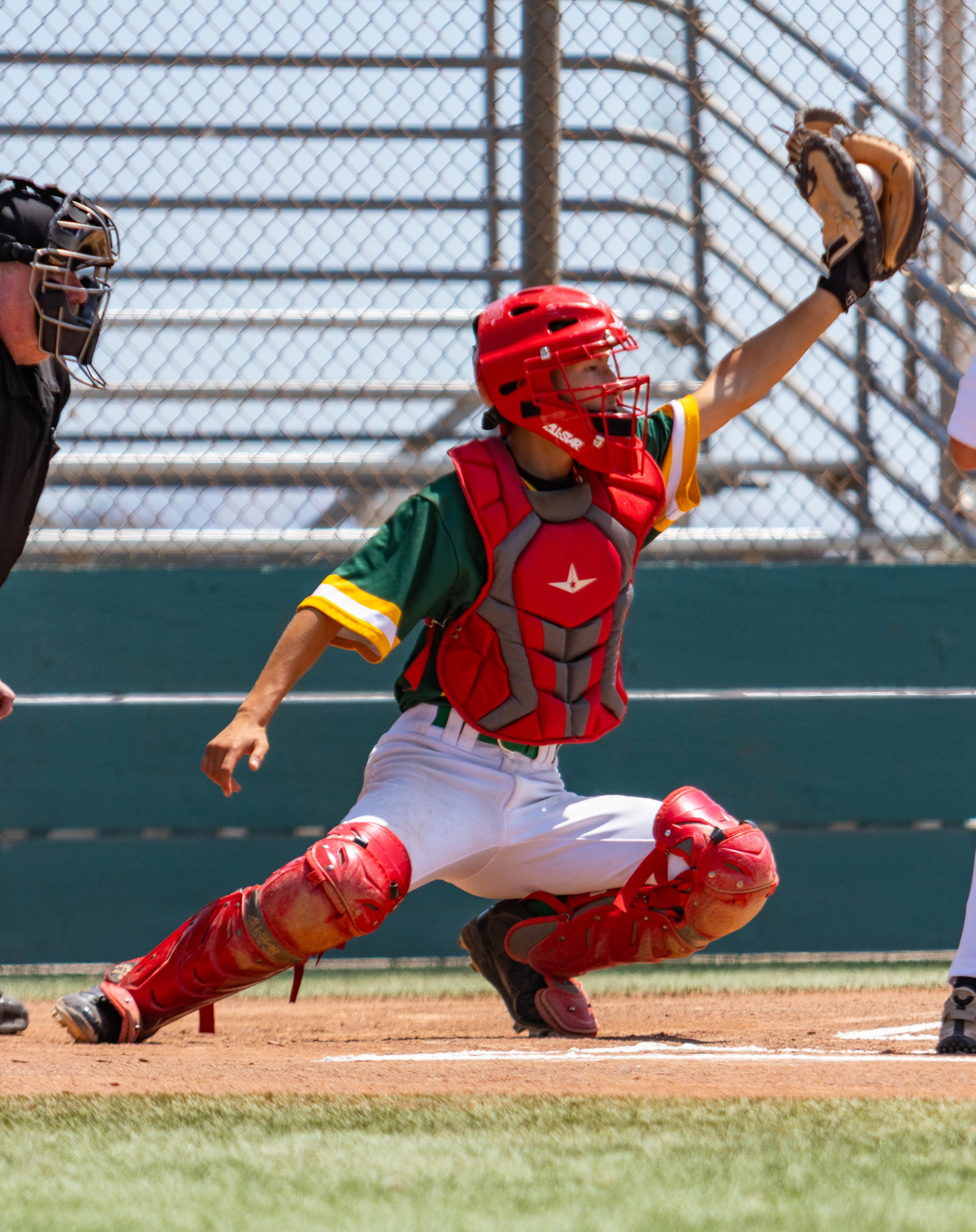 A female baseball catcher in a green uniform and red protective gear attempting to catch a ball during a game. The background features a chain-link fence and blue sky.
