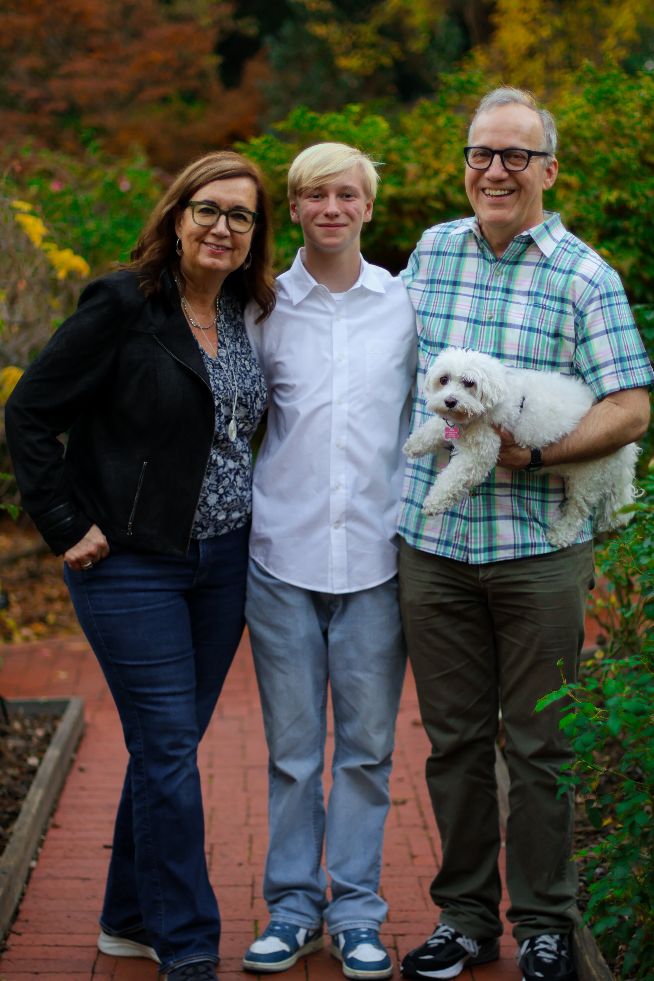 A family of three humans and a dog outdoors on a brick pathway with autumn foliage in the background. The woman on the left has dark hair, glasses, and is wearing a black jacket. The young boy in the middle has blond hair and is wearing a white shirt