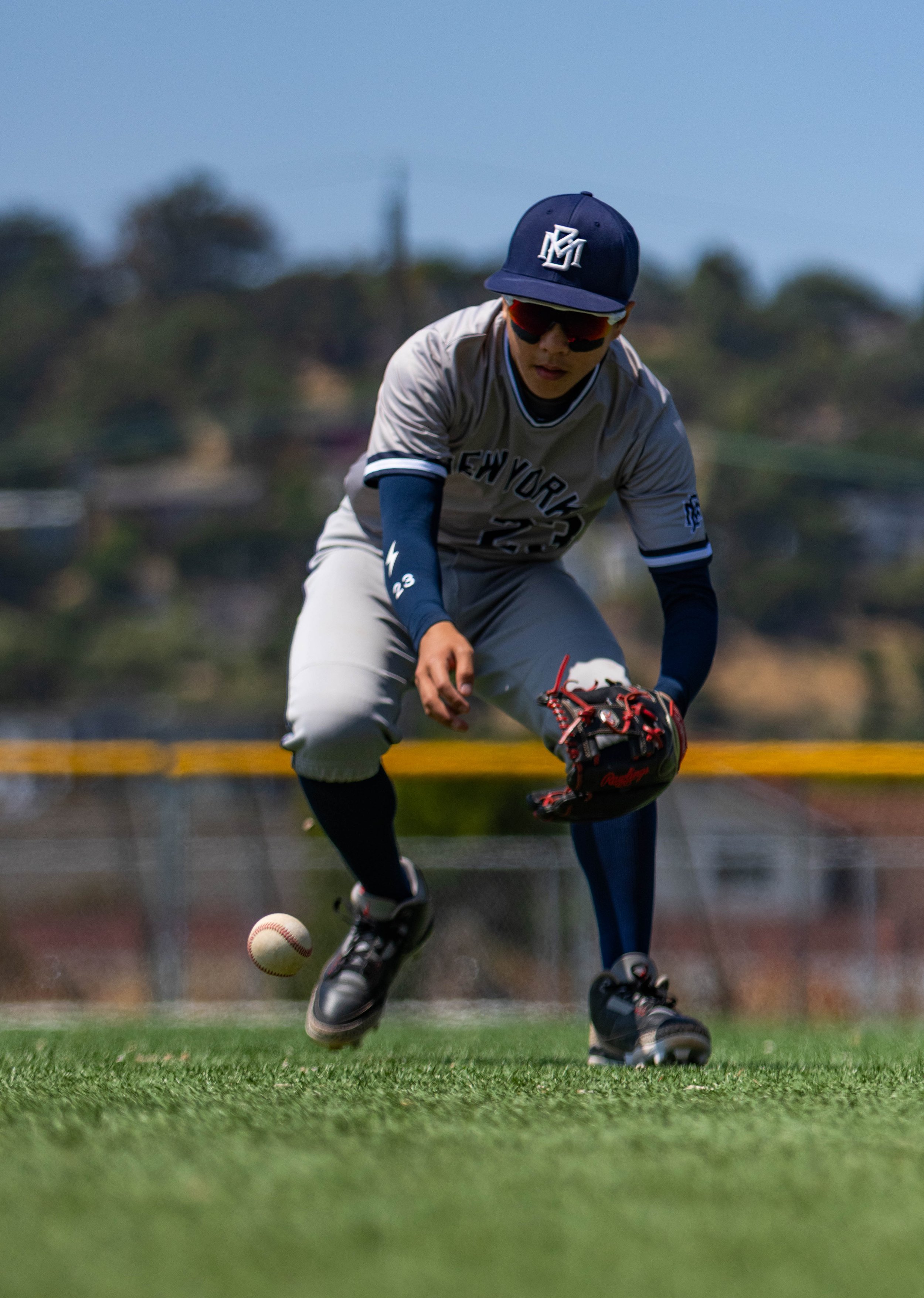 A baseball player in uniform and cap fields a ball on the field with a blurred background of trees and blue sky.