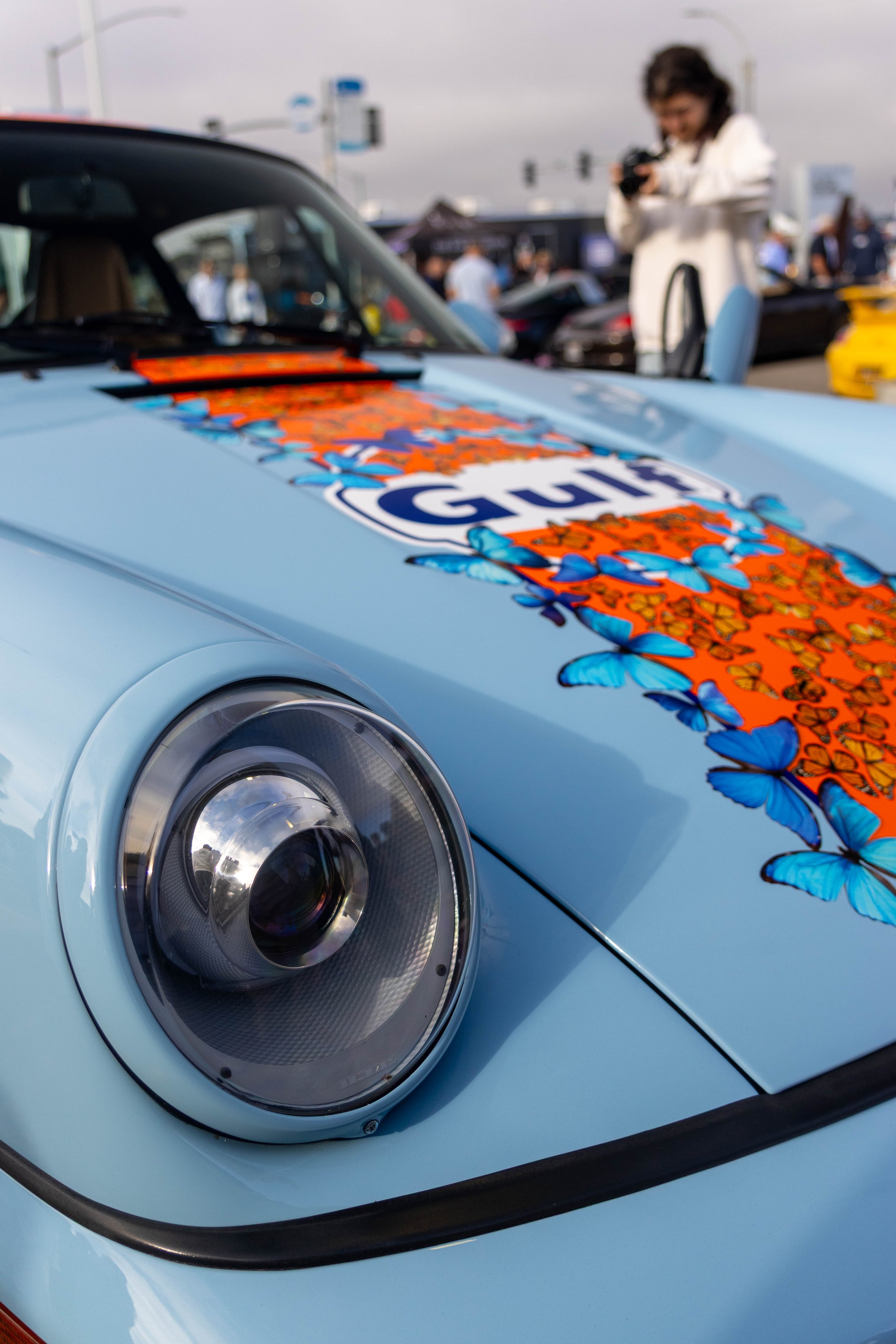 Close-up of a light blue sports car with a colorful butterfly decal and a partial Gulf logo on the hood, at an outdoor car show, with people in the background and a woman taking a photo.