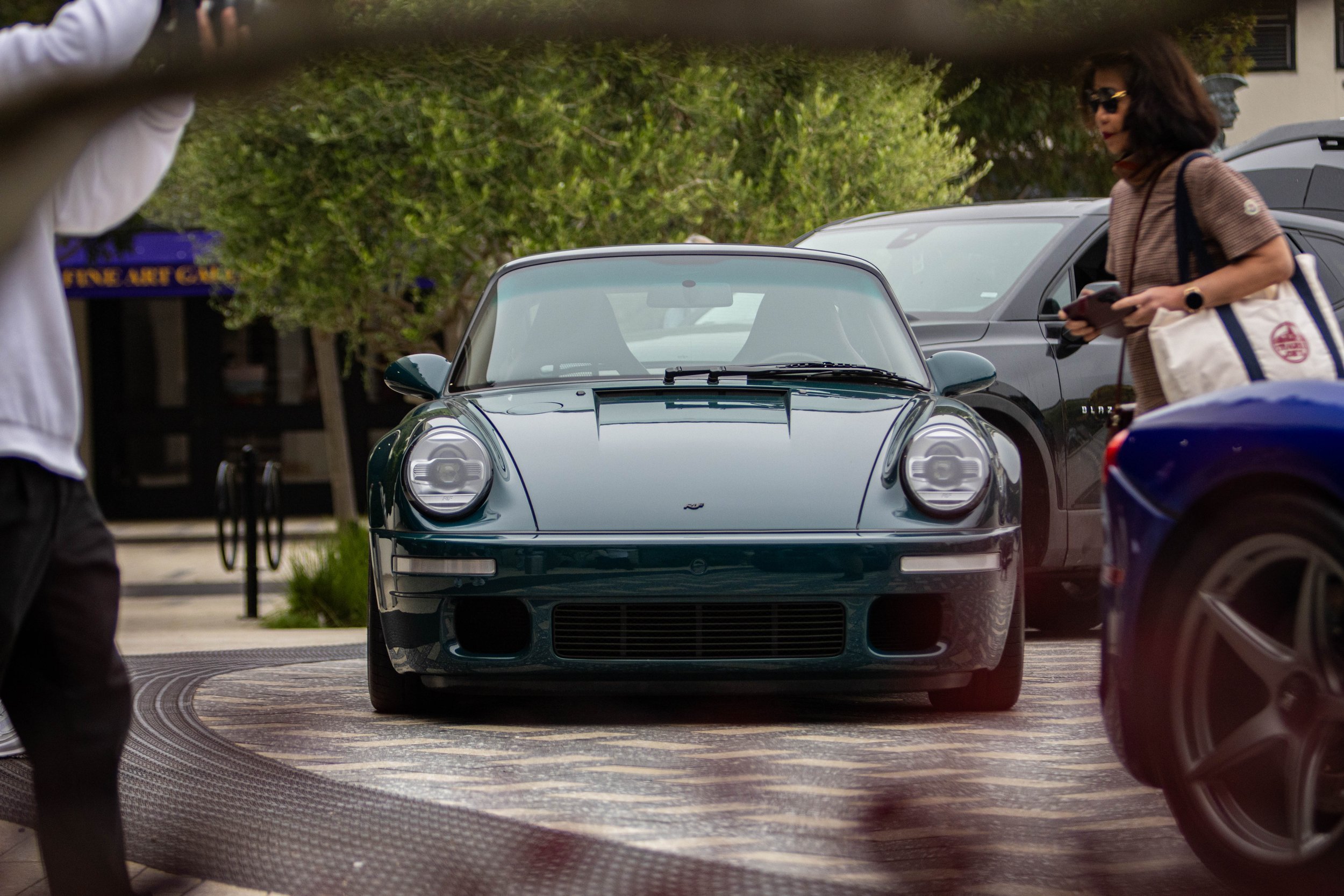 A woman with dark hair, wearing sunglasses, a brown shirt, and carrying a white bag, stands near a gray car and is looking at her phone in a parking lot with other cars and trees in the background.