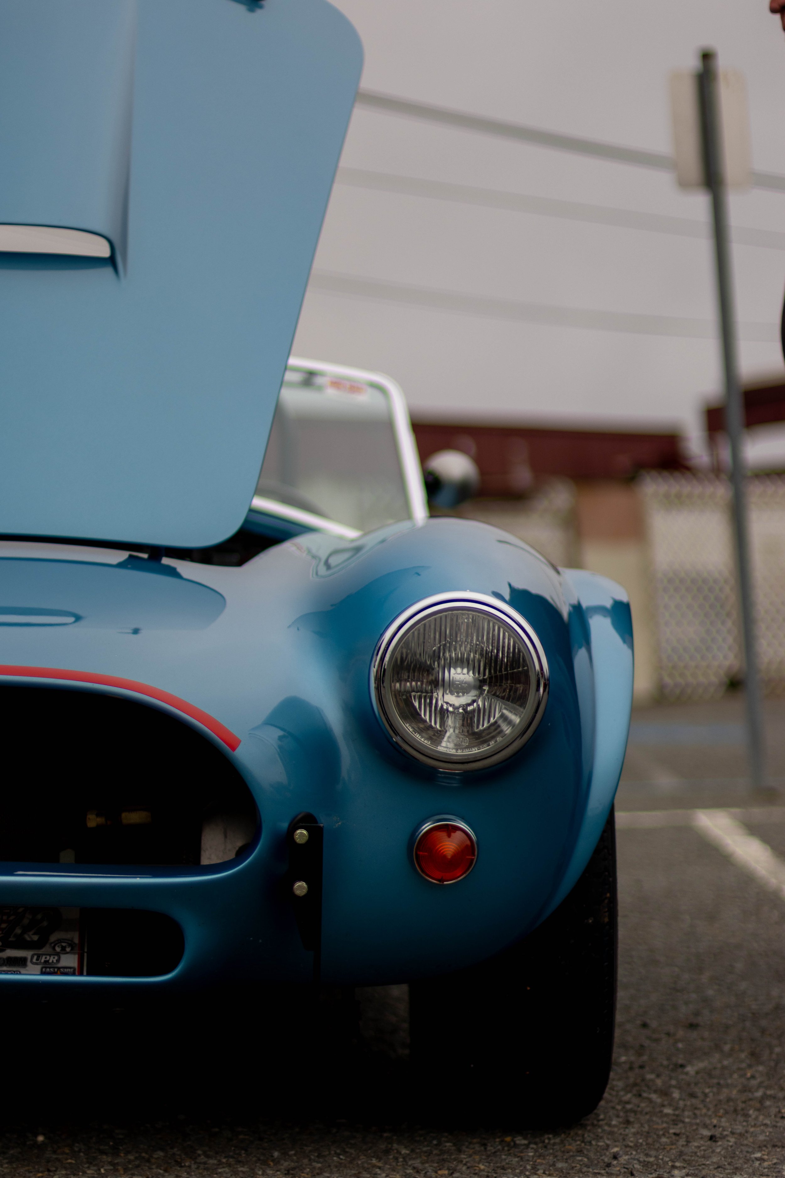 Close-up of front part of a blue vintage car with round headlight and orange turn signal, parked on asphalt with a background of a chain-link fence and a building.