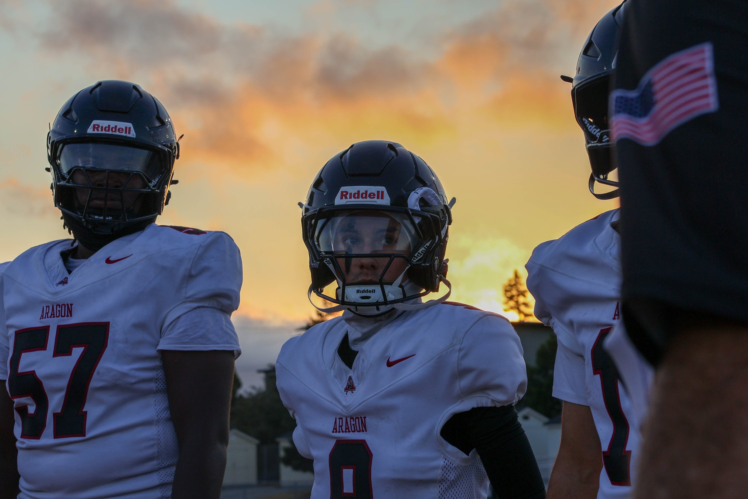Three American football players in white jerseys with red and black accents, helmets, and gear, standing outdoors during sunset.
