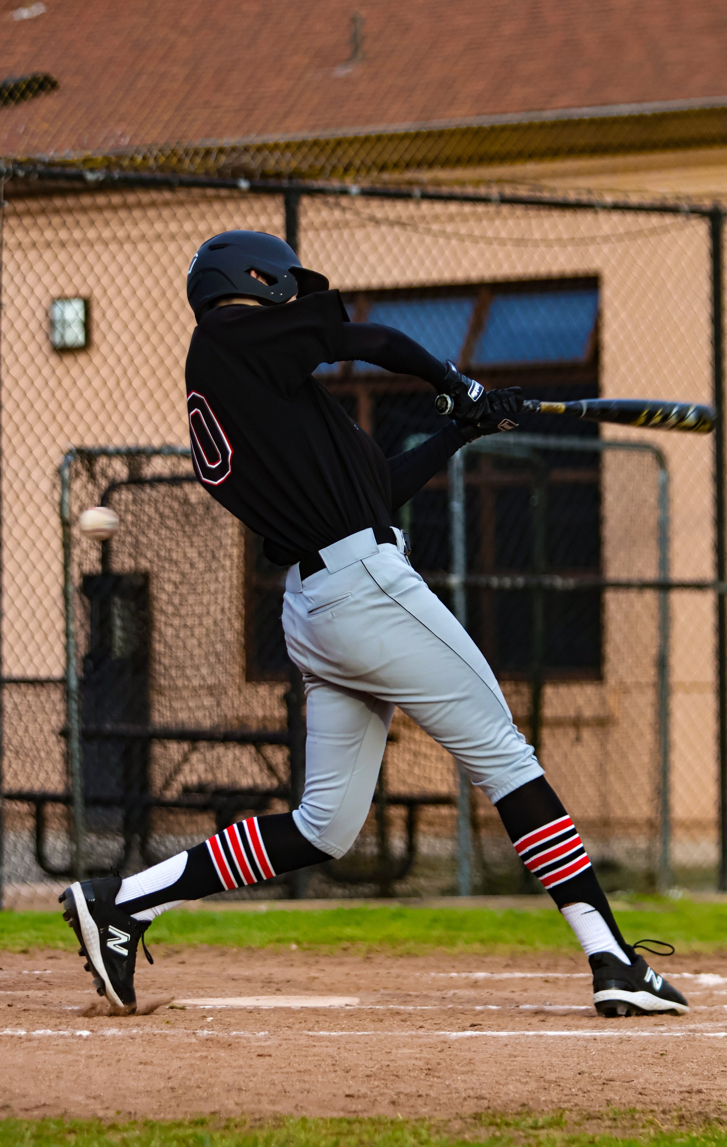 A baseball player batting during a game on a dirt field, wearing a black helmet, black jersey with the number 0, white pants, black and red striped socks, and black cleats.