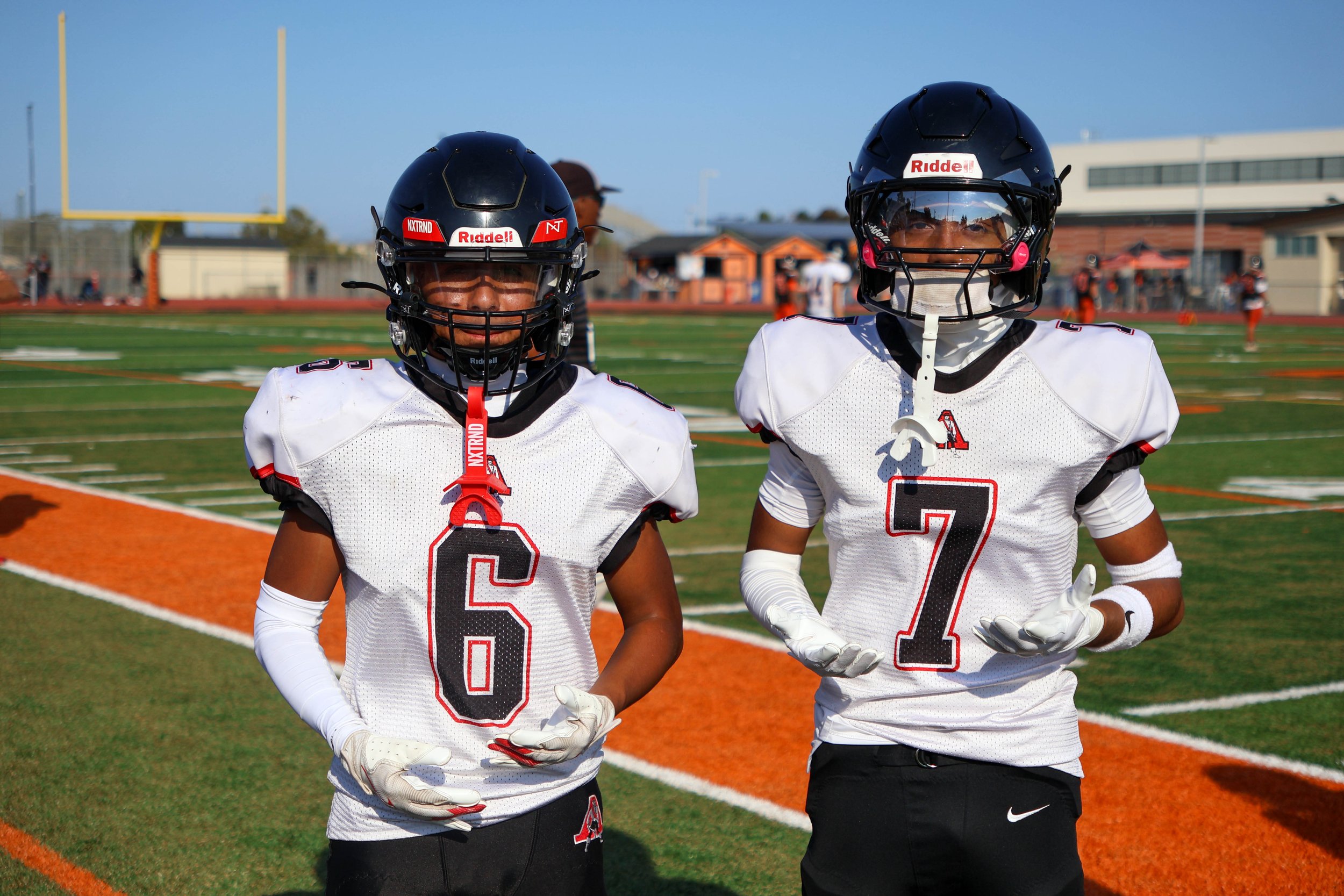 Two football players wearing black helmets and white jerseys with black and red details standing on a football field with a goalpost in the background.