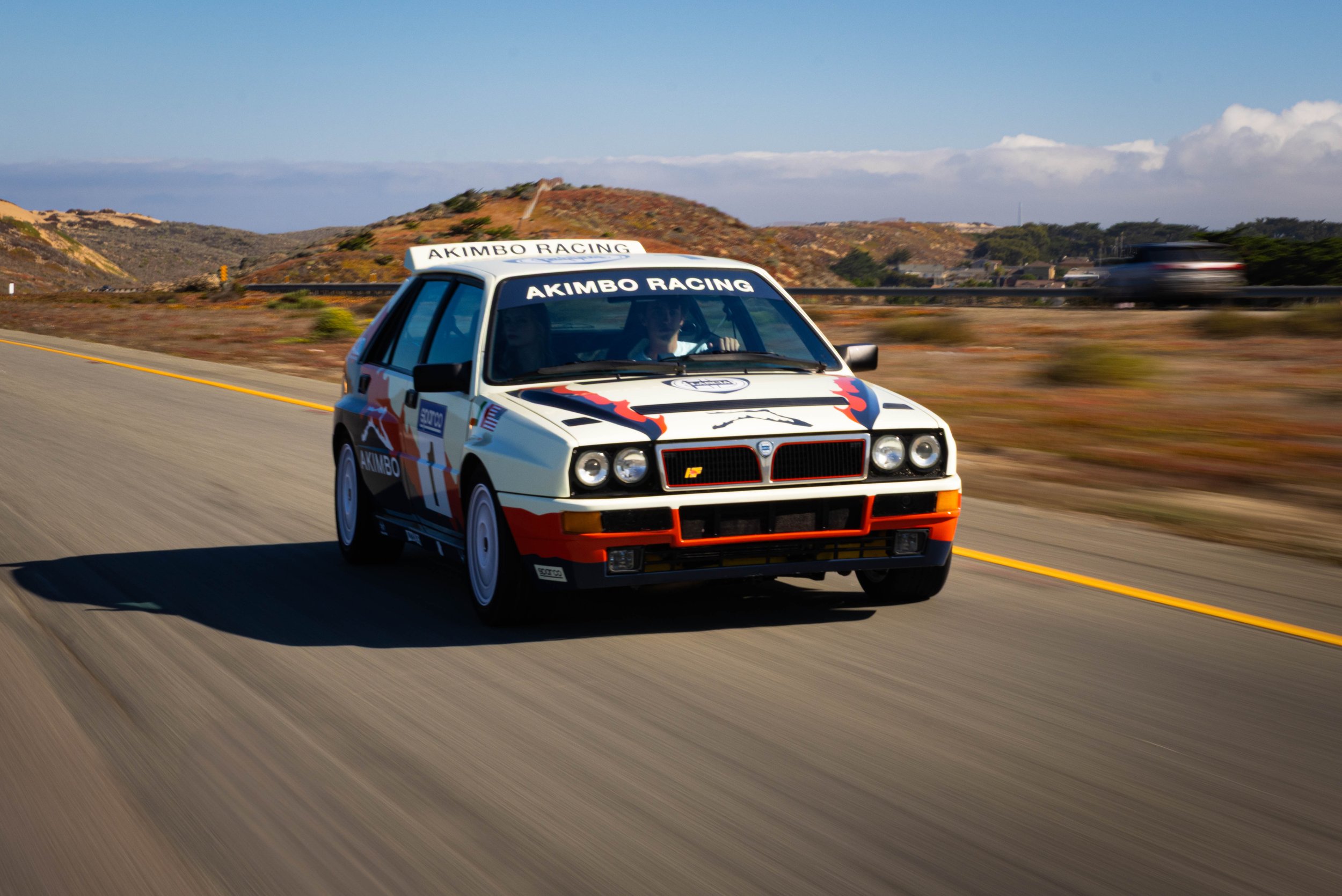 A vintage white race car with colorful branding, driving on an open highway through a desert landscape with rolling hills and a blue sky.