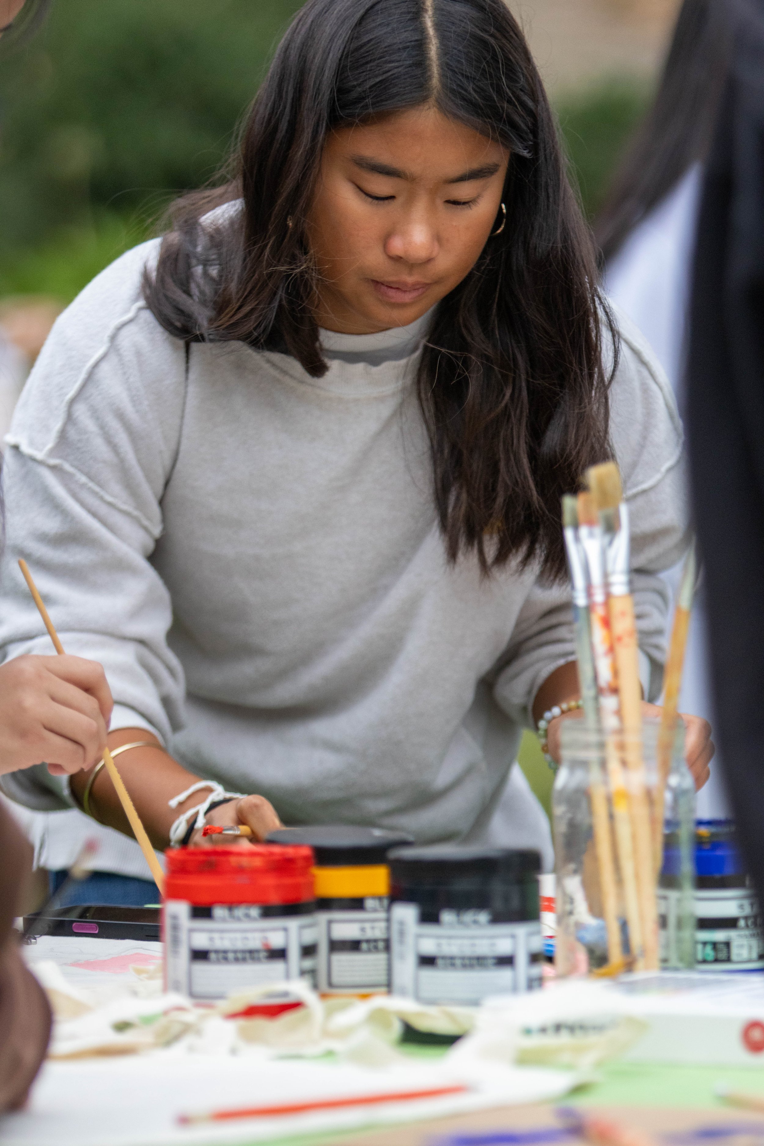 A young woman with long dark hair and hoop earrings painting on a canvas outdoors, with art supplies and paint bottles on the table in front of her.