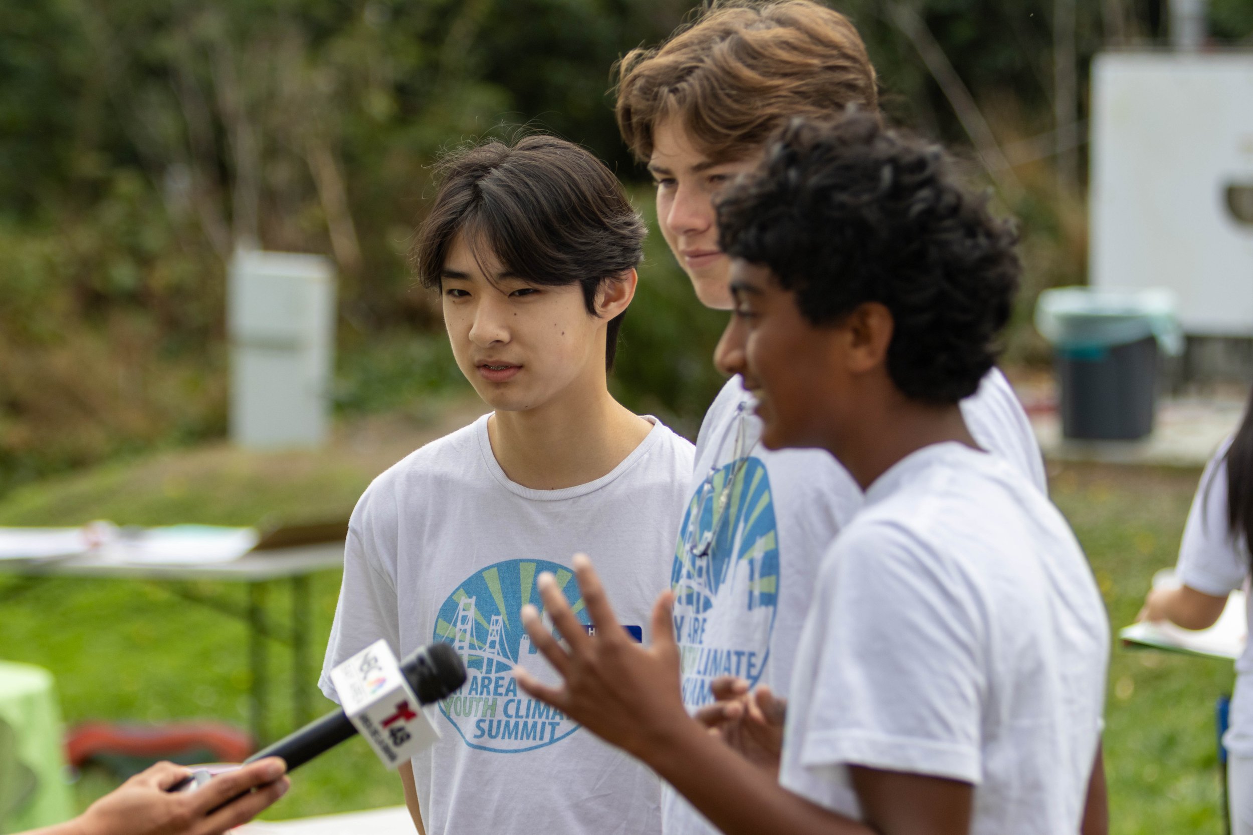 Group of young people outdoors at a climate summit, wearing white T-shirts with a climate-themed logo, engaging in conversation with a microphone and reporters.