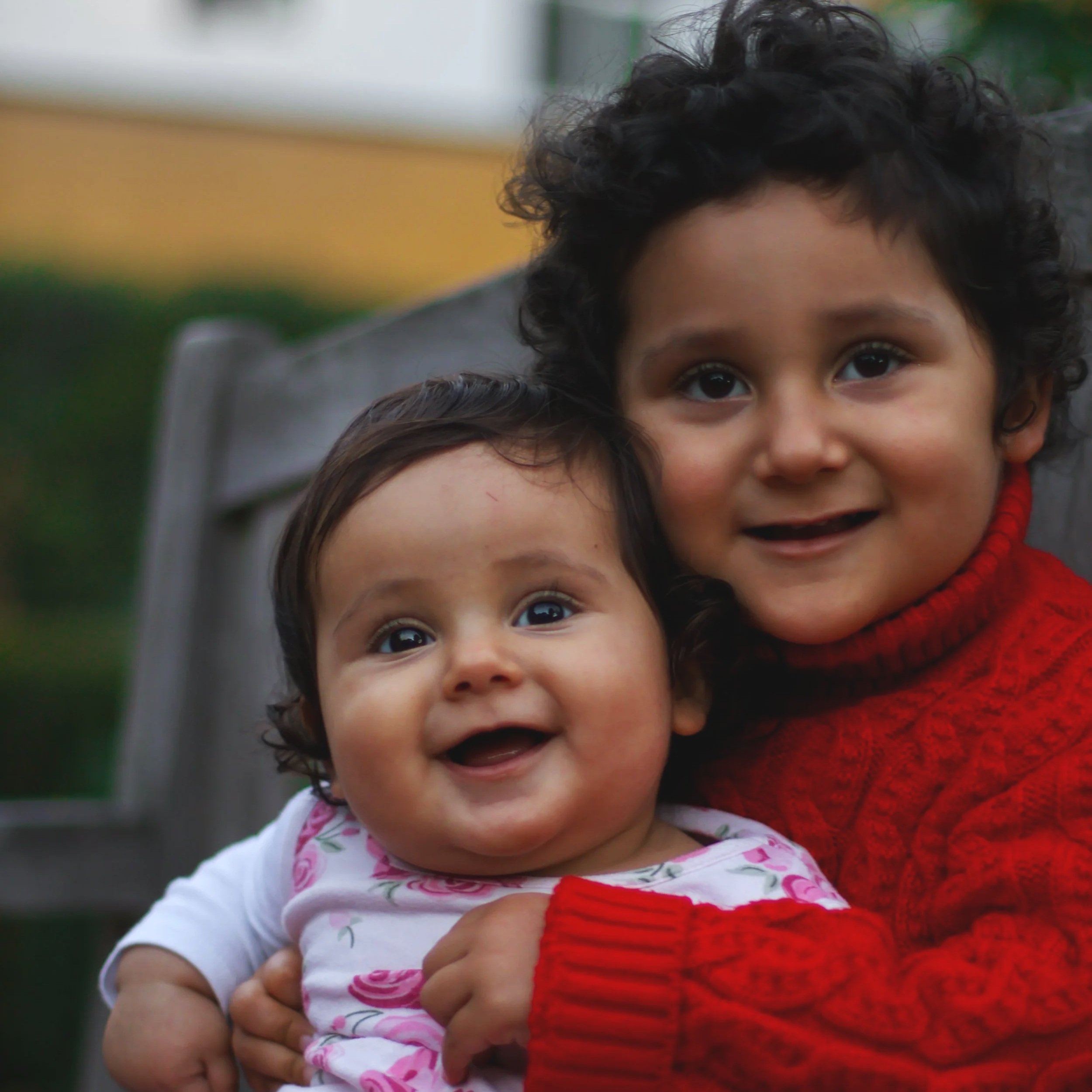 Close-up of two children, a young girl with curly black hair and a baby, possibly her sibling, with big brown eyes and dark hair, smiling outdoors.