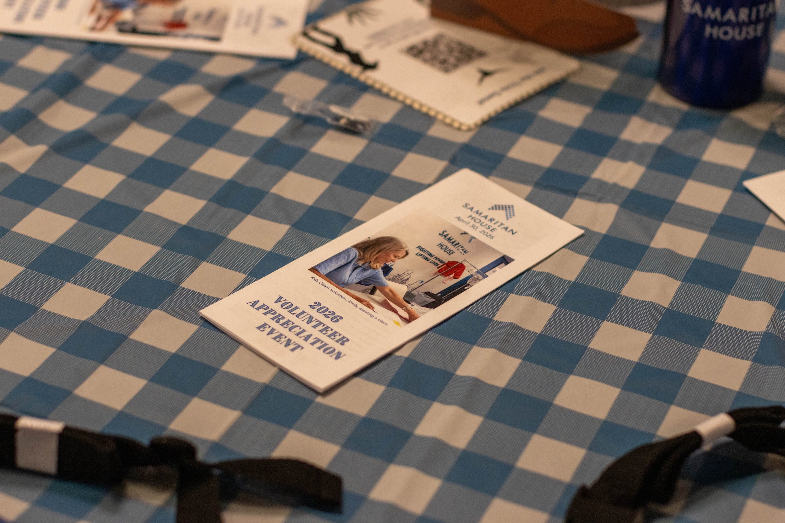 A table covered with a blue and white checkered tablecloth holding flyers and informational materials for a volunteer appreciation event at Samaritan House.