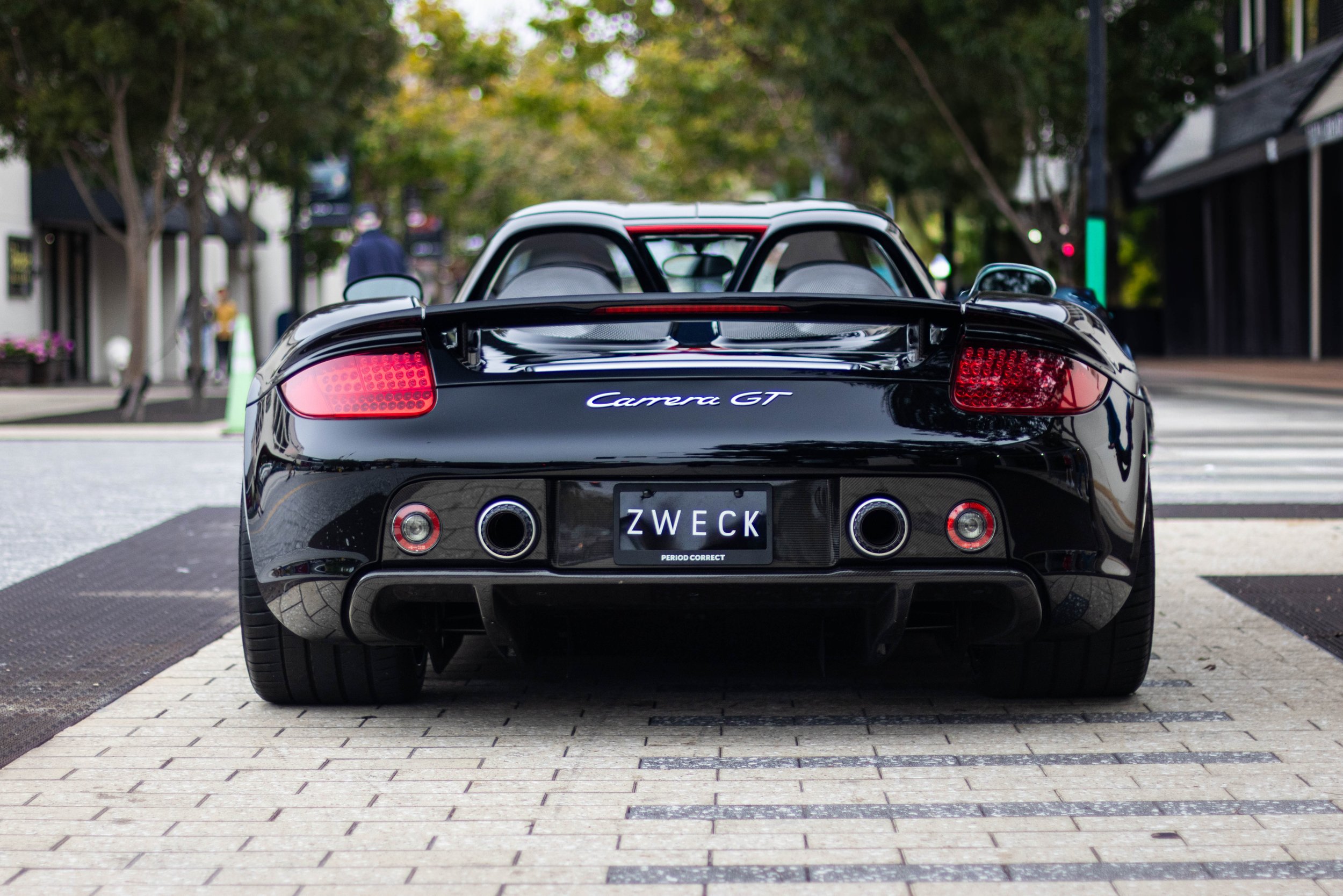Back view of a black Porsche Carrera GT sports car parked on a city street with trees and buildings in the background.