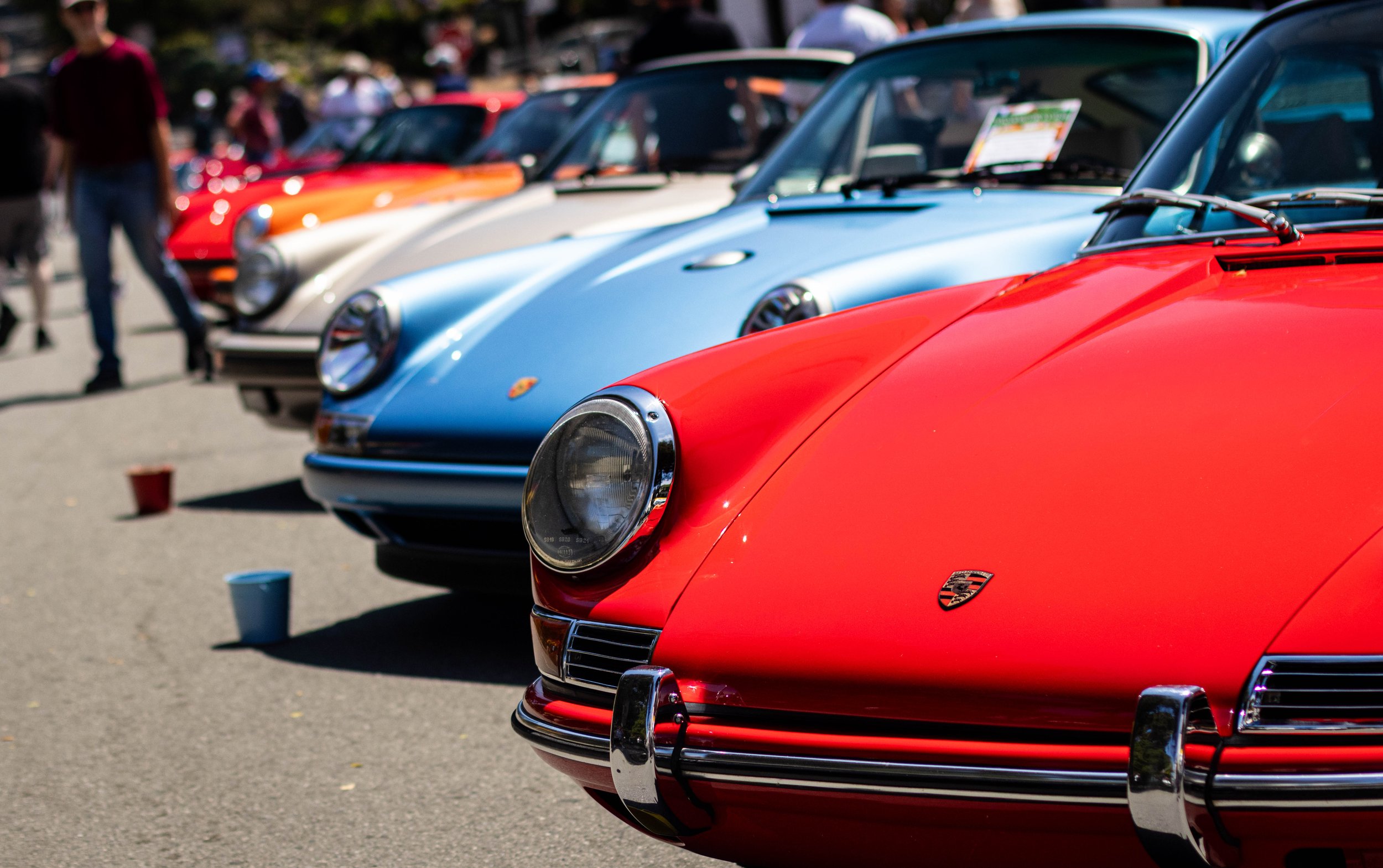 Lineup of colorful classic Porsche cars at a car show, with people walking in the background.