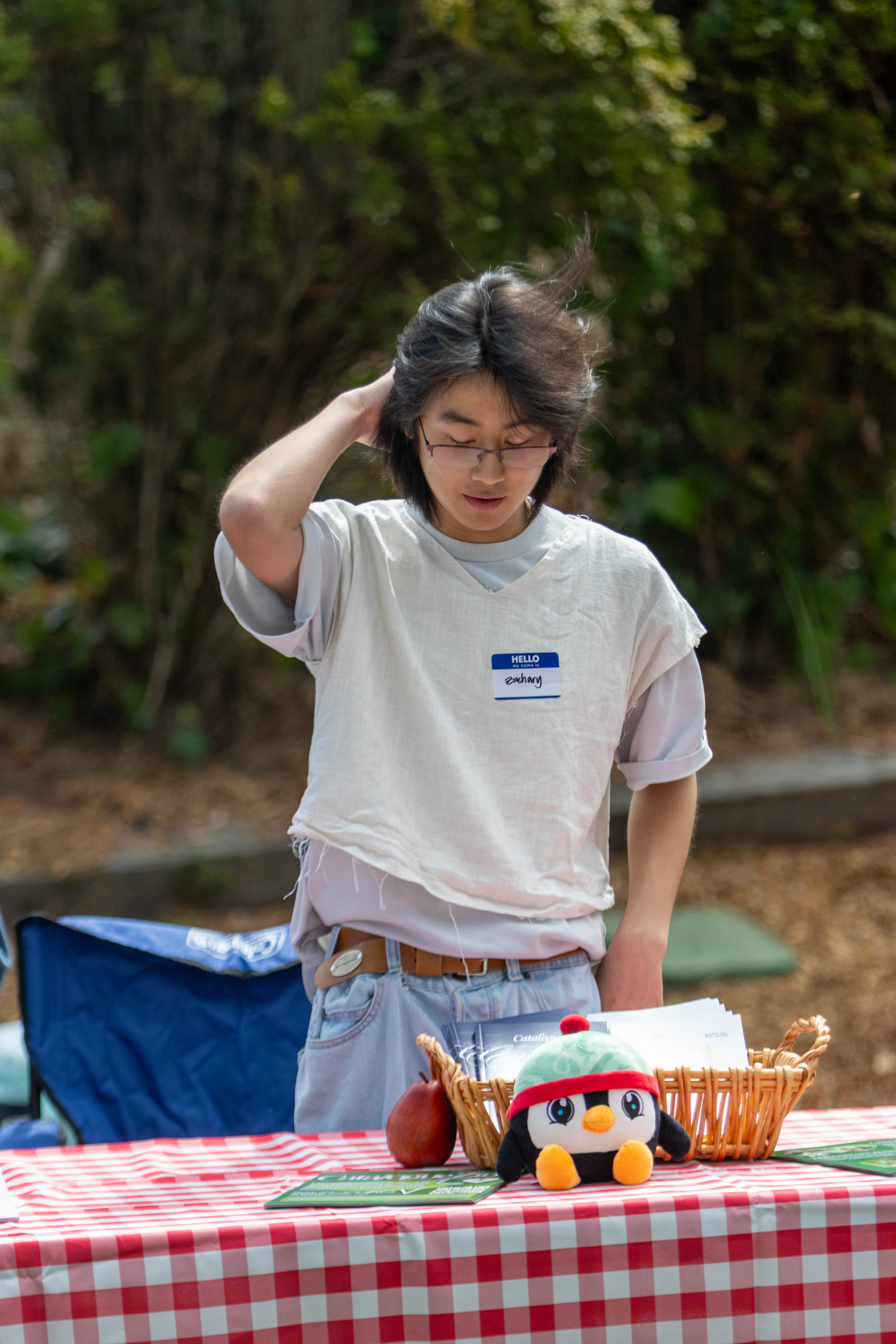 A person with glasses and a name tag that says 'Zachary' is standing behind a table with a red and white checkered tablecloth. The table has some papers, a small stuffed penguin, an apple, and a basket with some items on it. The person is outdoors wi