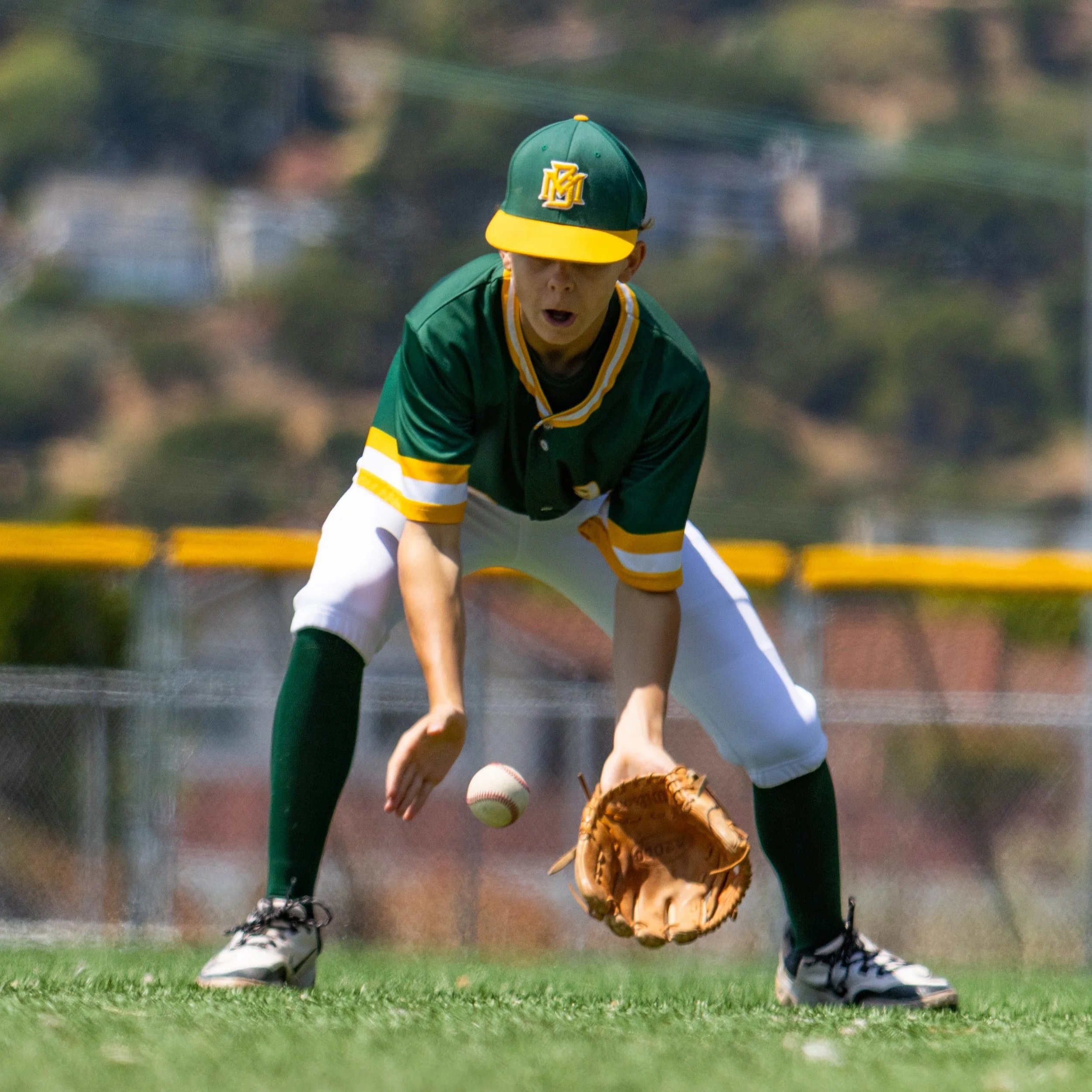 A baseball player in green and yellow uniform and cap, kneeling on a field, with a person in white pants and shoes on the ground nearby.