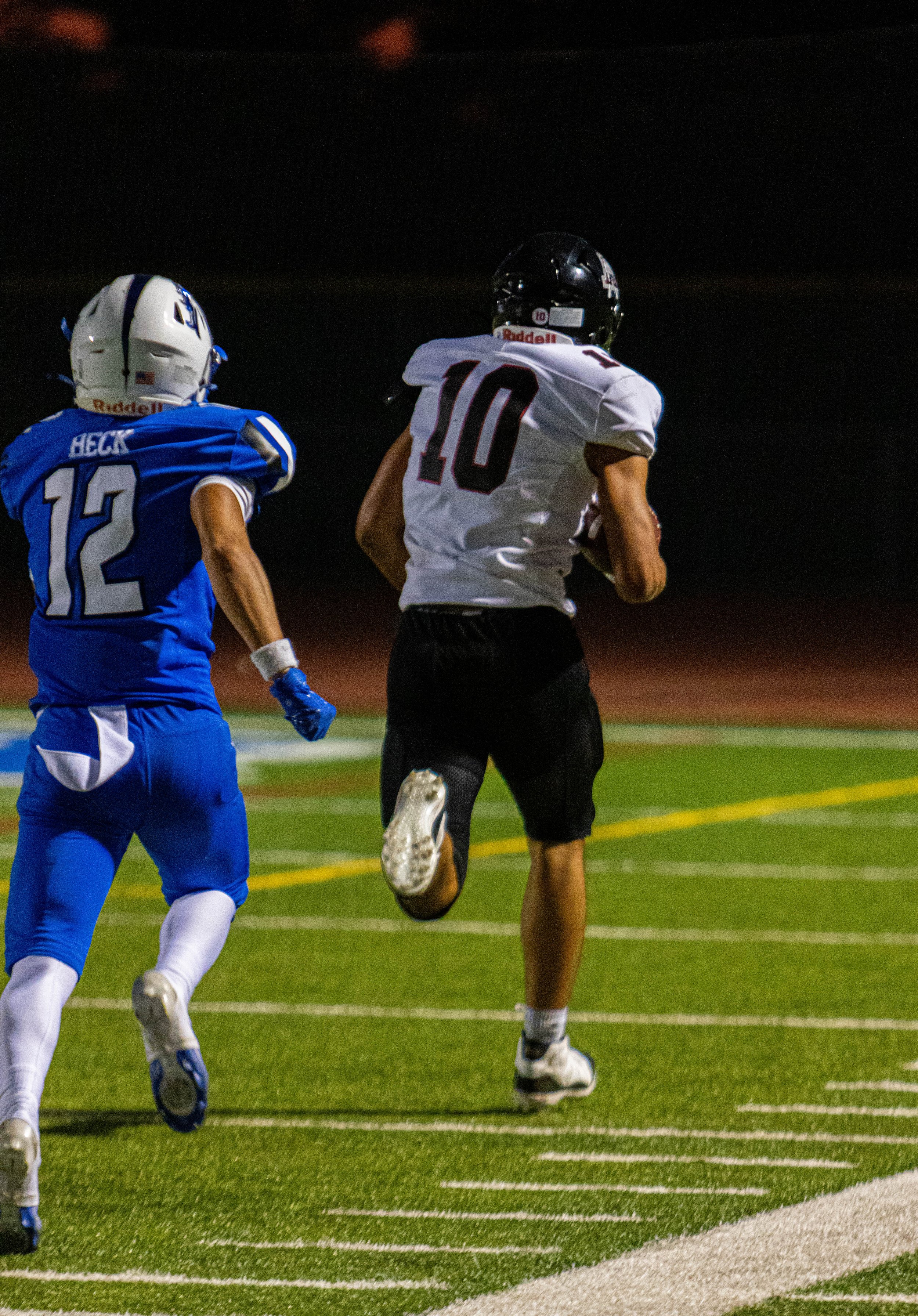 A nighttime scene of two high school football players running on the sideline of a football field, with one in a white uniform and black helmet with the number 10, and the other in a blue uniform and white helmet with the number 12.