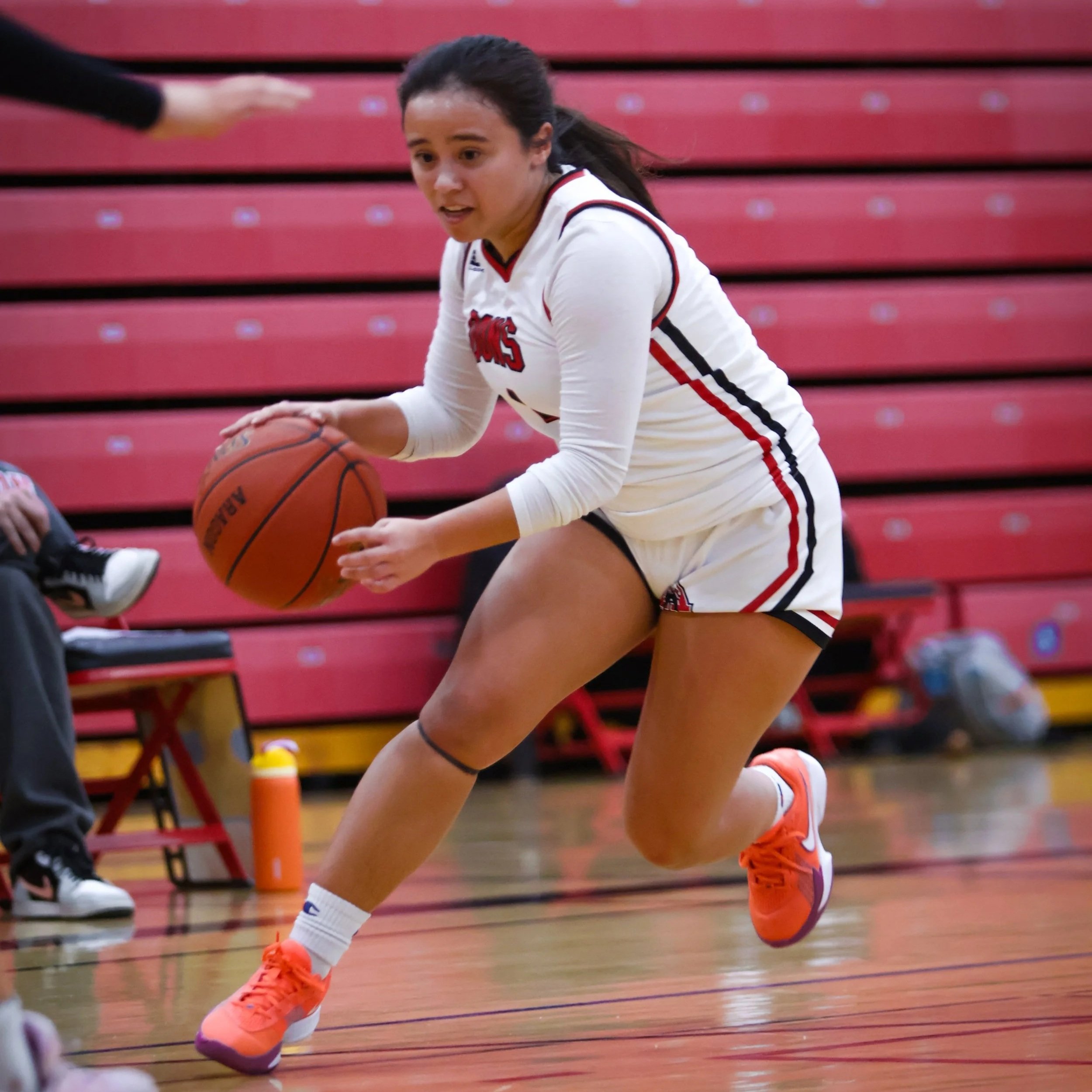 Female basketball player in white uniform with red and black accents, orange shoes, and white socks, crouched on a basketball court handling an orange basketball with black stripes, with red bleachers in the background.