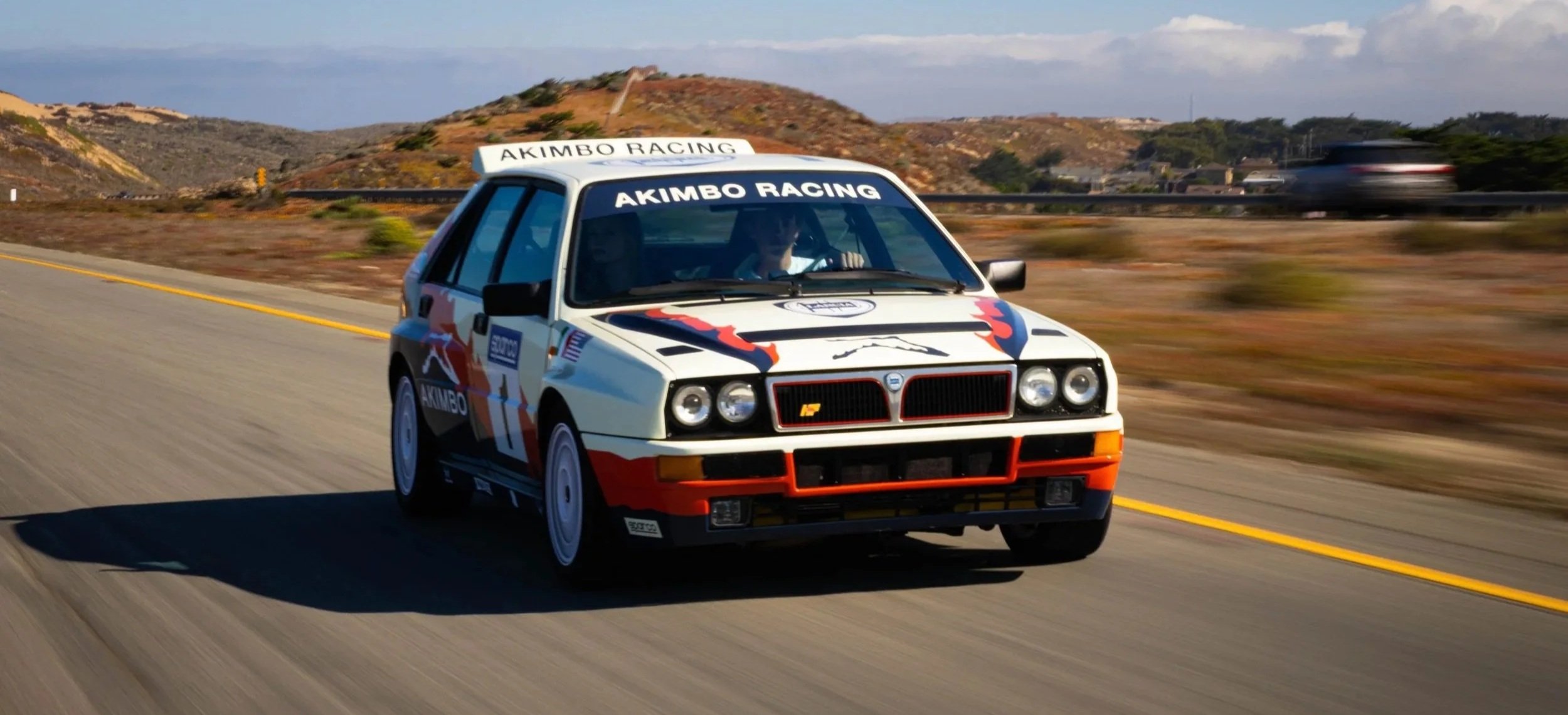 A rally built Alfa Romeo with red, black, and blue graphics and the words 'AKIMBO RACING' on the windshield is driving on a highway through a desert landscape. The background features hills, a guardrail, and a blurred passing vehicle.