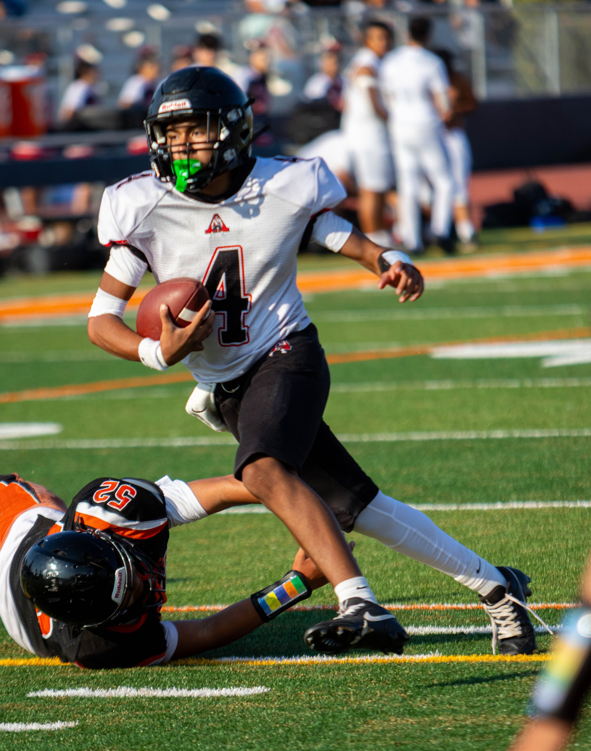 A young football player wearing a white jersey with the number 4, black helmet with faceguard and a green mouthguard, holding a football and running on the field. Another player, wearing a black and orange helmet, is on the ground in front of him. In