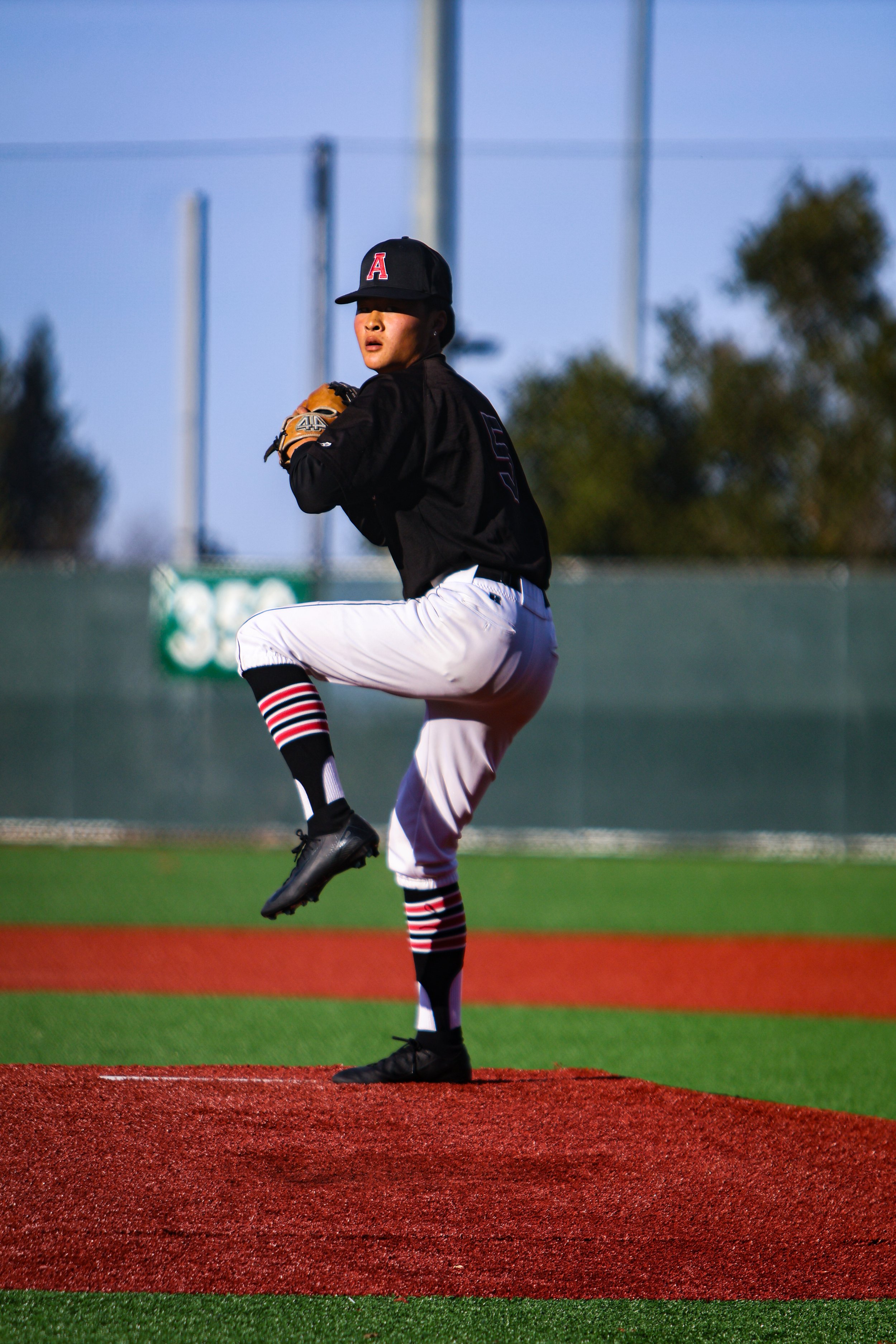 A baseball player is pitching on the mound during a game, wearing a black cap with a red A, black jersey, and white pants. The scene is outdoors with a blue sky and blurred background of trees and outfield.