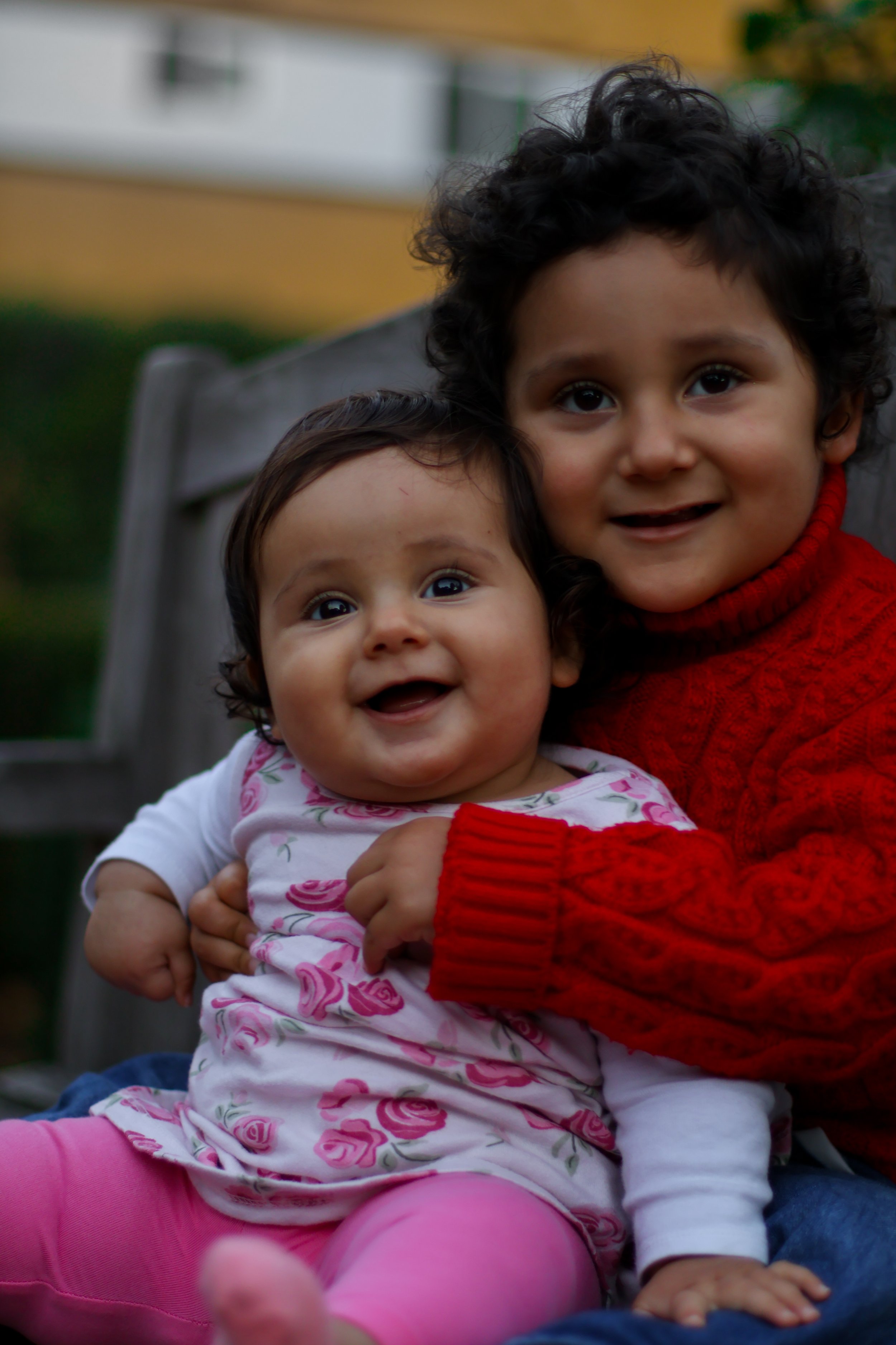 Two young children, a girl and a boy, are sitting together outdoors on a wooden bench. The girl has curly dark hair and is wearing a pink dress with a floral pattern, pink leggings, and a long-sleeved white shirt. The boy, with dark, curly hair, is w