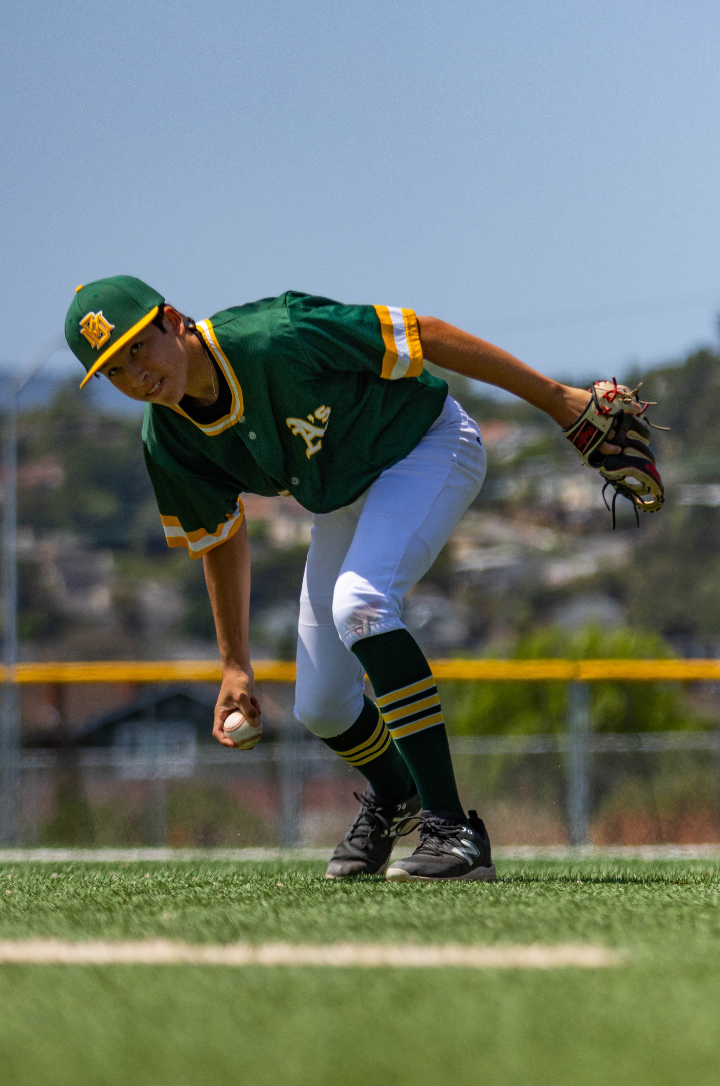 A young male baseball player in a green uniform with yellow and white accents, wearing a green cap with yellow trim and a logo, is bending down on a baseball field to pick up a baseball, with a glove on his left hand. The background features a blue s