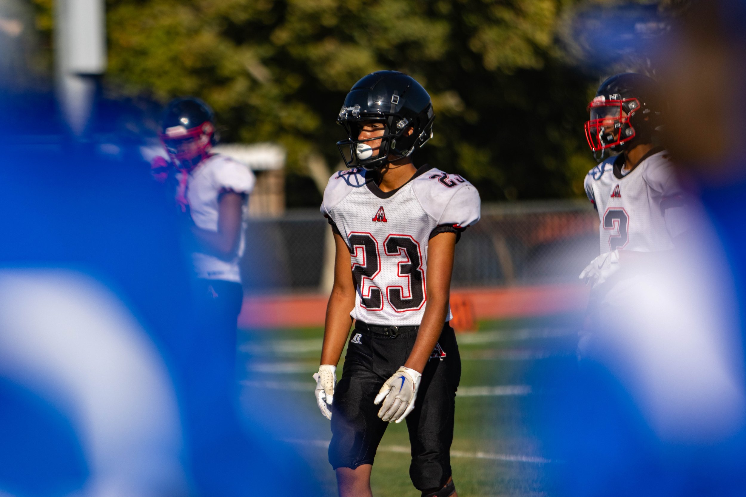 A football player wearing a white jersey with black and red accents, black pants, gloves, and a black helmet stands on a football field. The player is facing slightly to the right, with other football players blurred in the background.