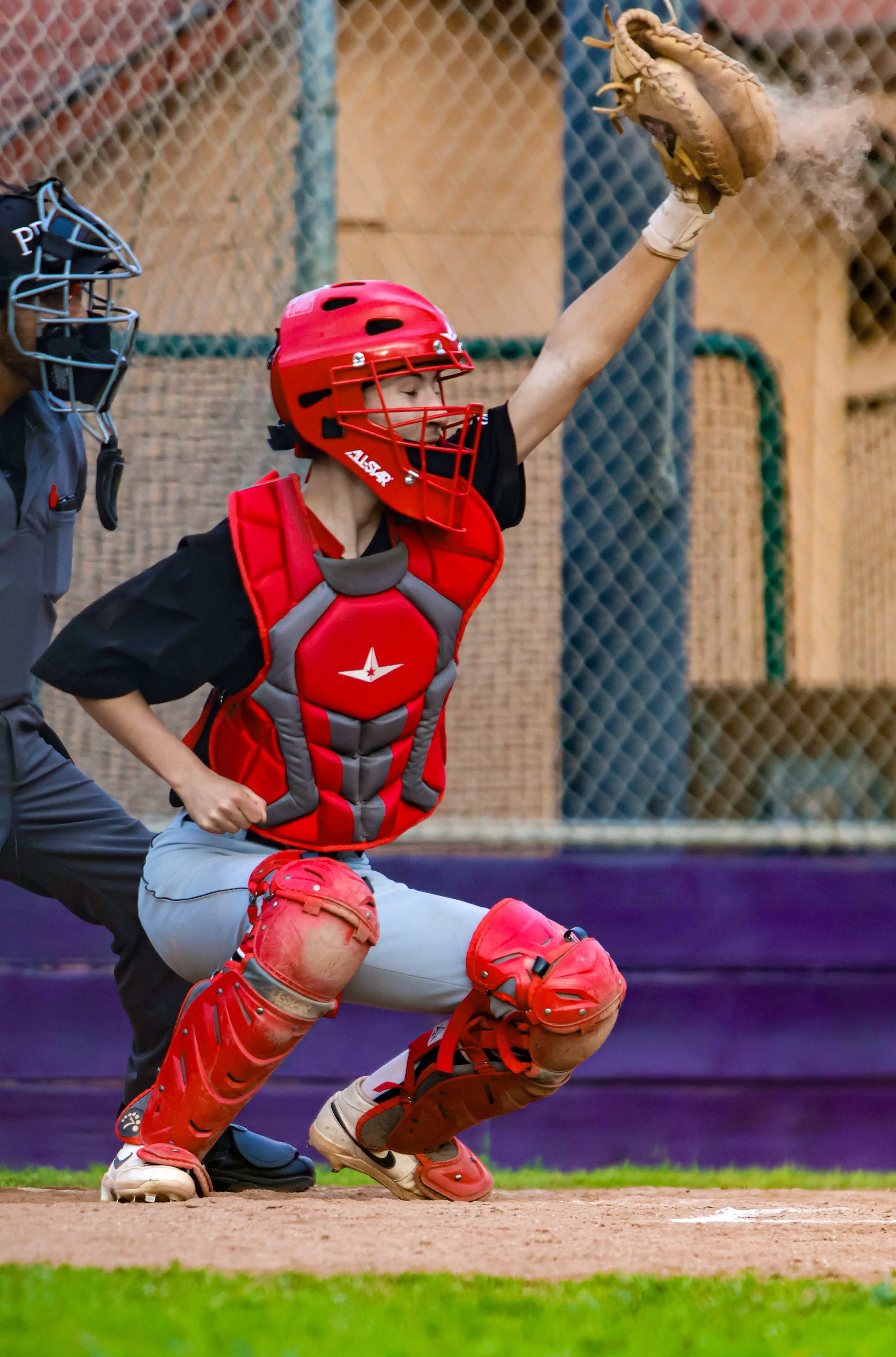 A young baseball catcher in a red helmet and padding, crouching behind home plate with a coach or umpire standing next to him during a game.