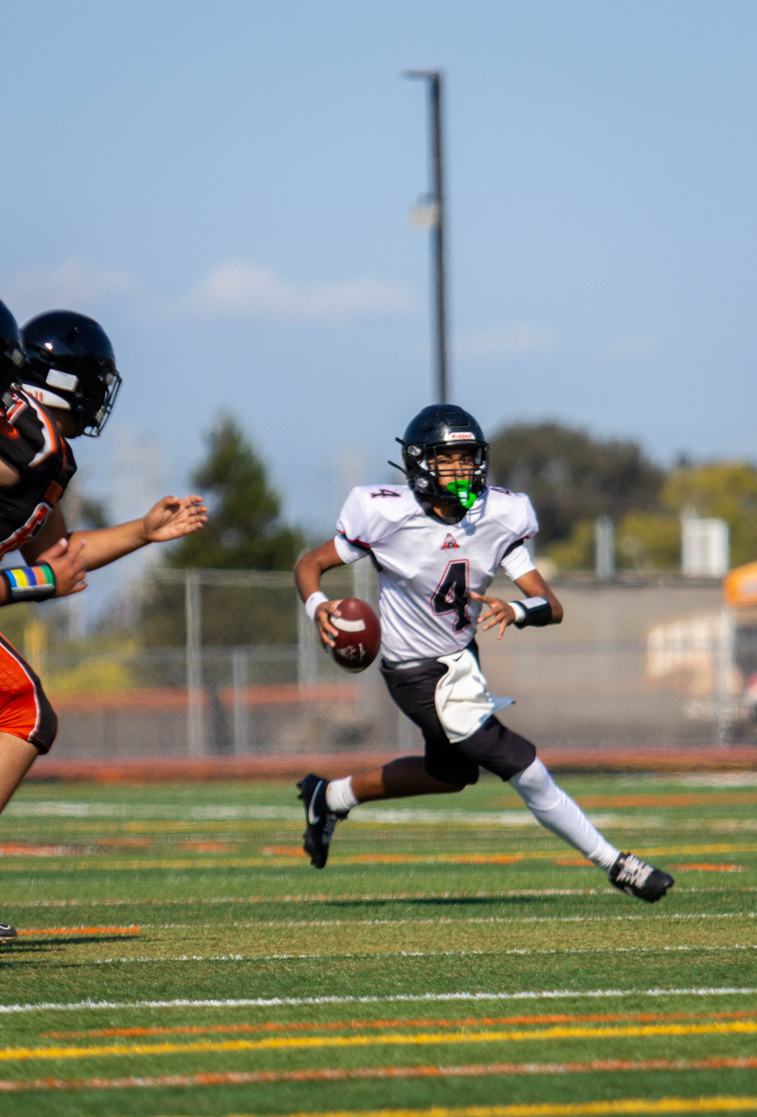 A youth football player wearing a white jersey with a red number 4, black pants, and a black helmet is running on a football field while holding a football. Other players are visible in the background, and the sky is clear.