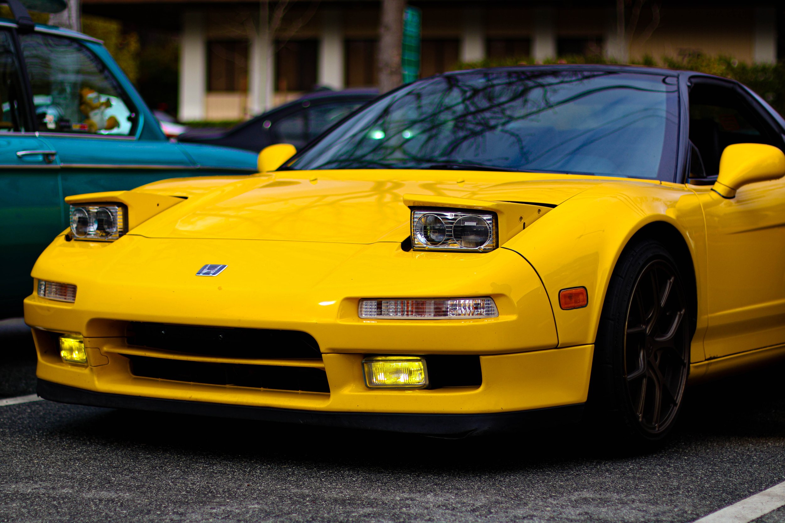 Close-up of a bright yellow sports car with black-tinted windows parked in a lot, with a green vintage car and other vehicles in the background.