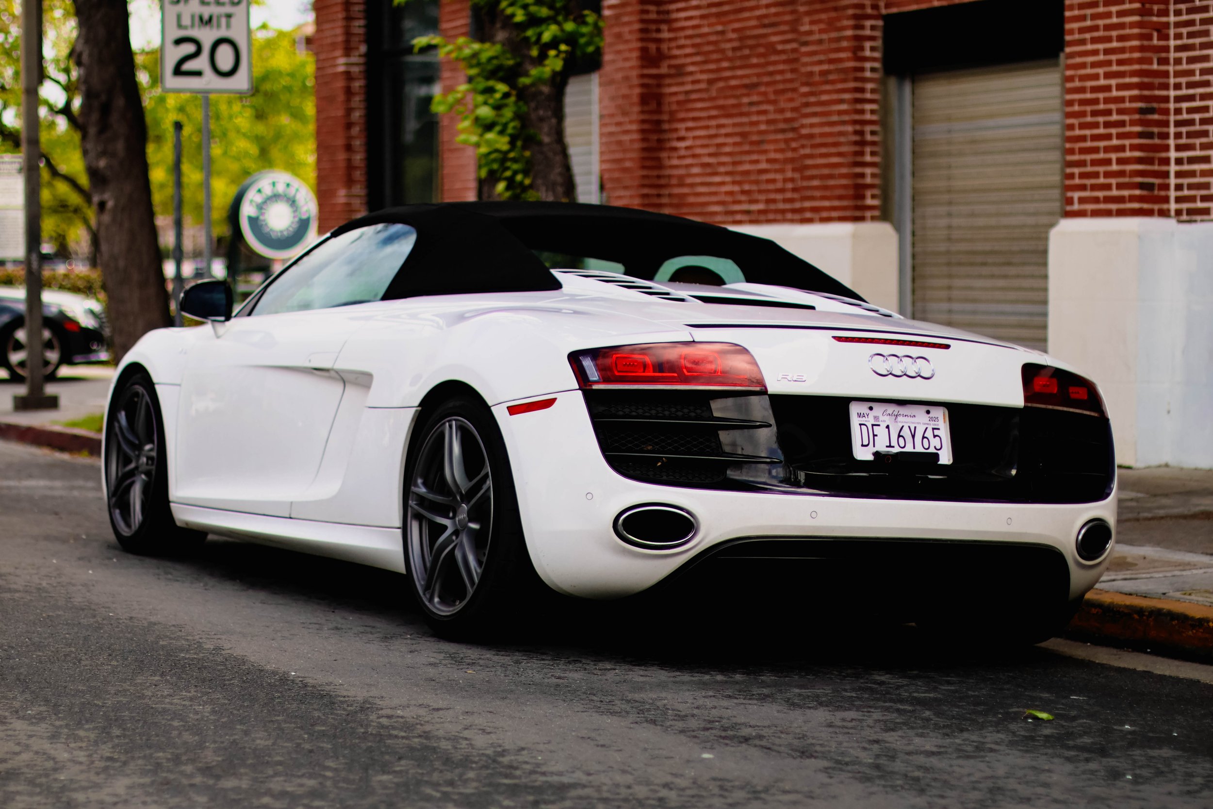 White convertible sports car with a black soft top parked on the street in front of a brick building and a green sign indicating a speed limit of 20 miles per hour.