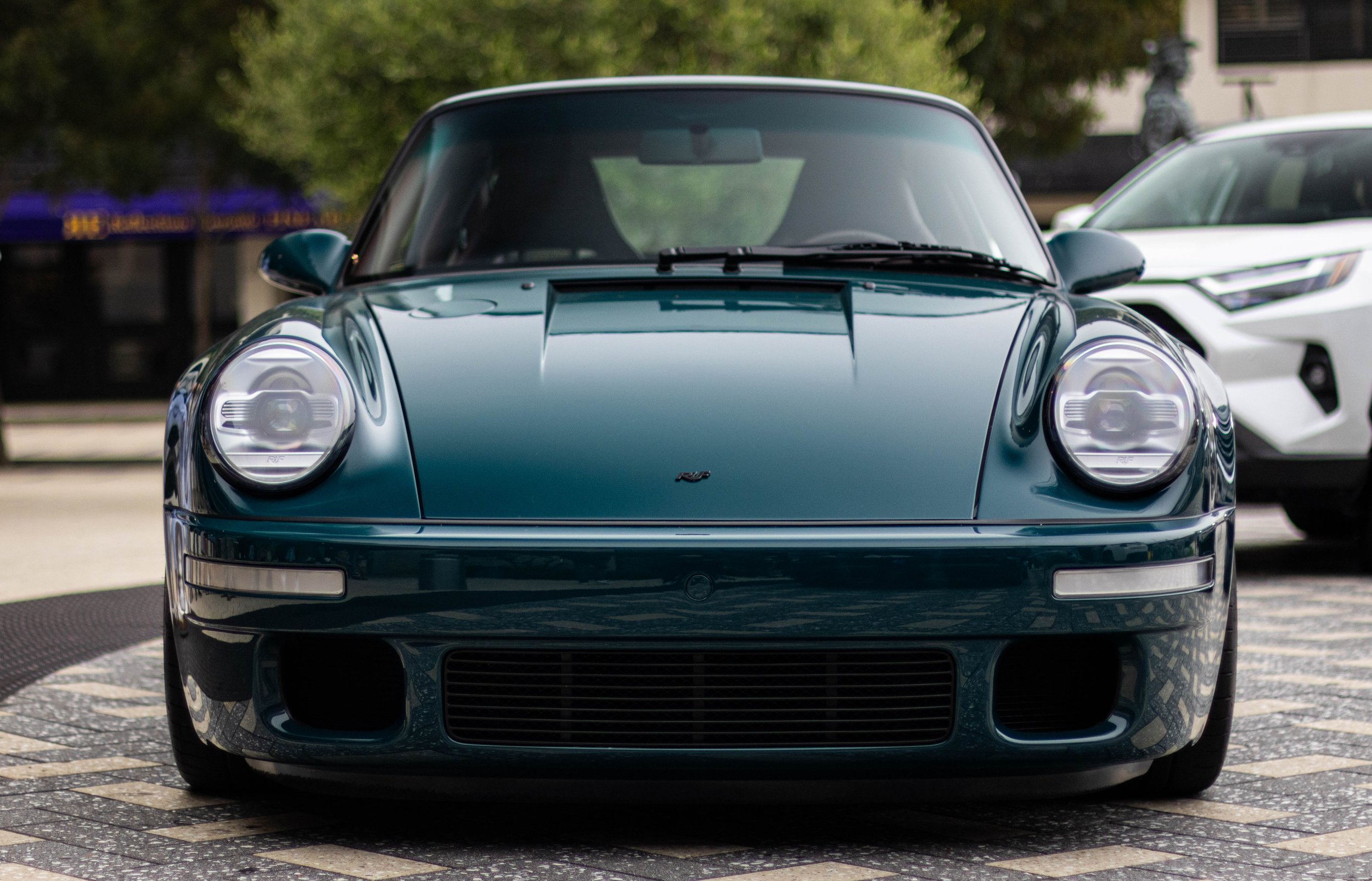 Front view of a black Porsche sports car parked on a brick-patterned surface with other cars and trees in the background.