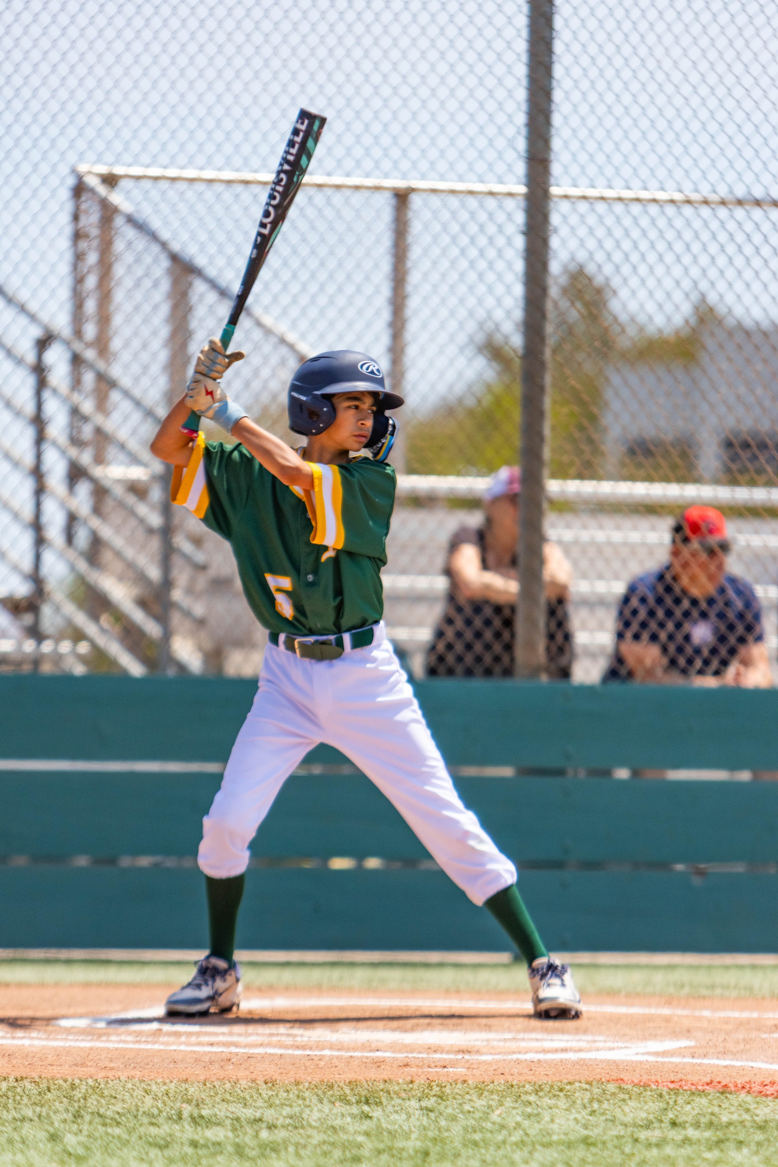 A young baseball player in a green uniform and helmet preparing to hit a ball at home plate in a baseball field.